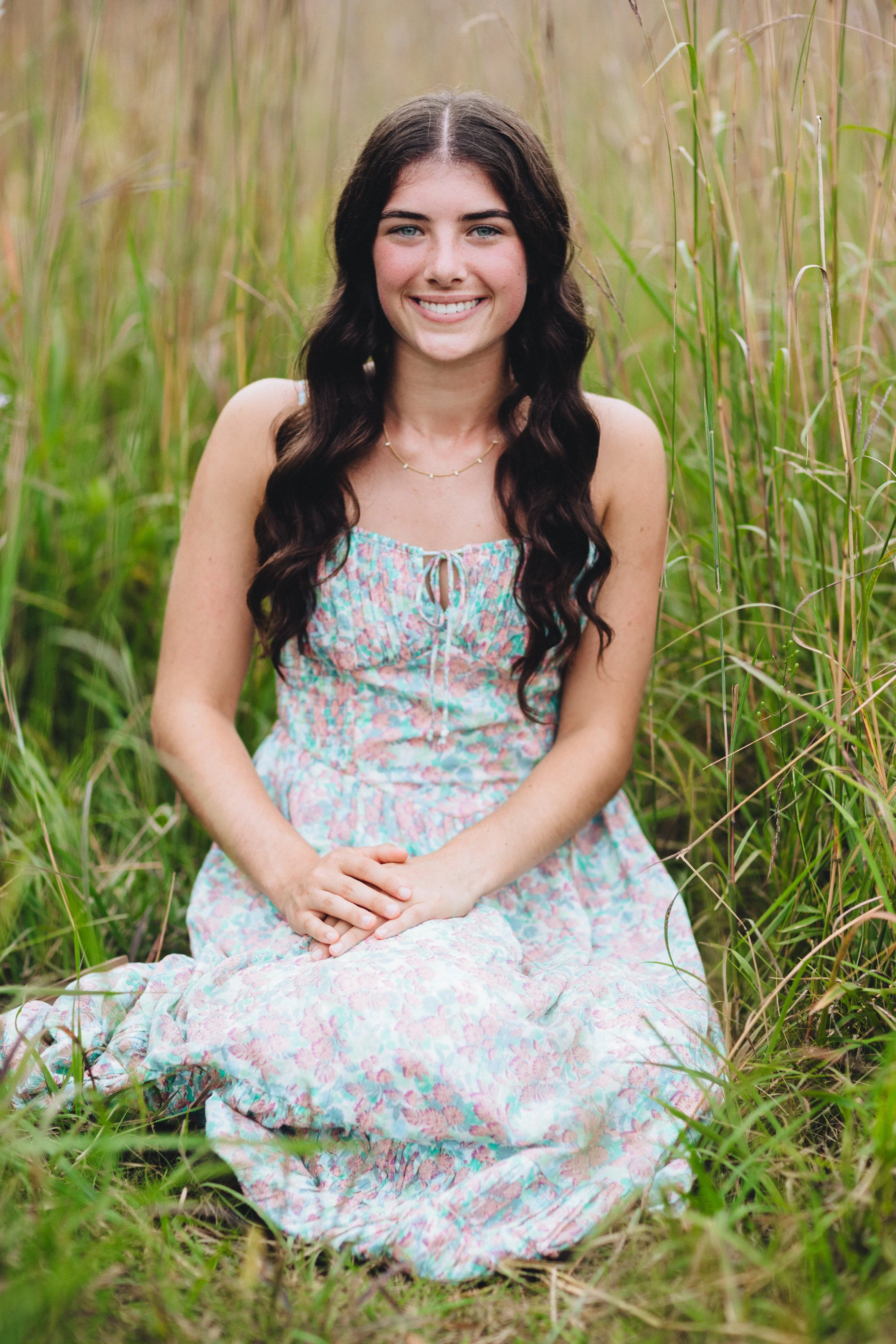 A young woman with long dark wavy hair, blue eyes, and a bright smile sitting on the grass among tall green grass in a natural outdoor setting, wearing a pastel-colored floral summer dress and a delicate necklace.