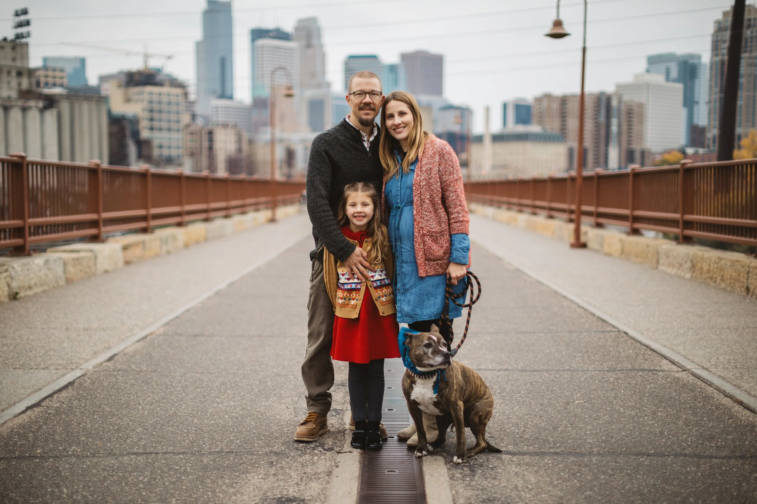 Family of four with dog posing on a city bridge with tall buildings in the background.