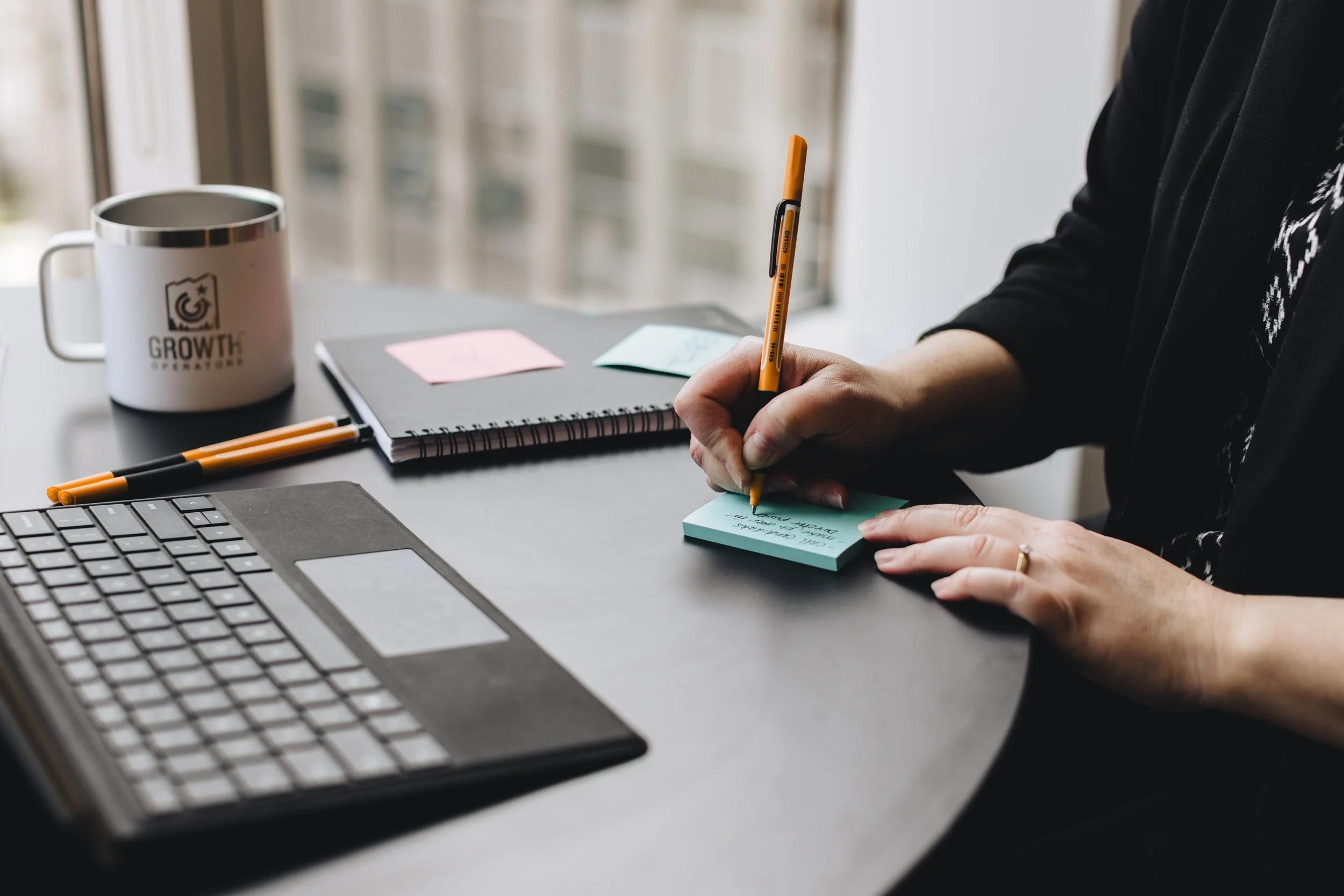 Person writing notes on sticky notes at a desk with a laptop, notebook, pens, and a coffee mug that says 'GROWTH' near a window.