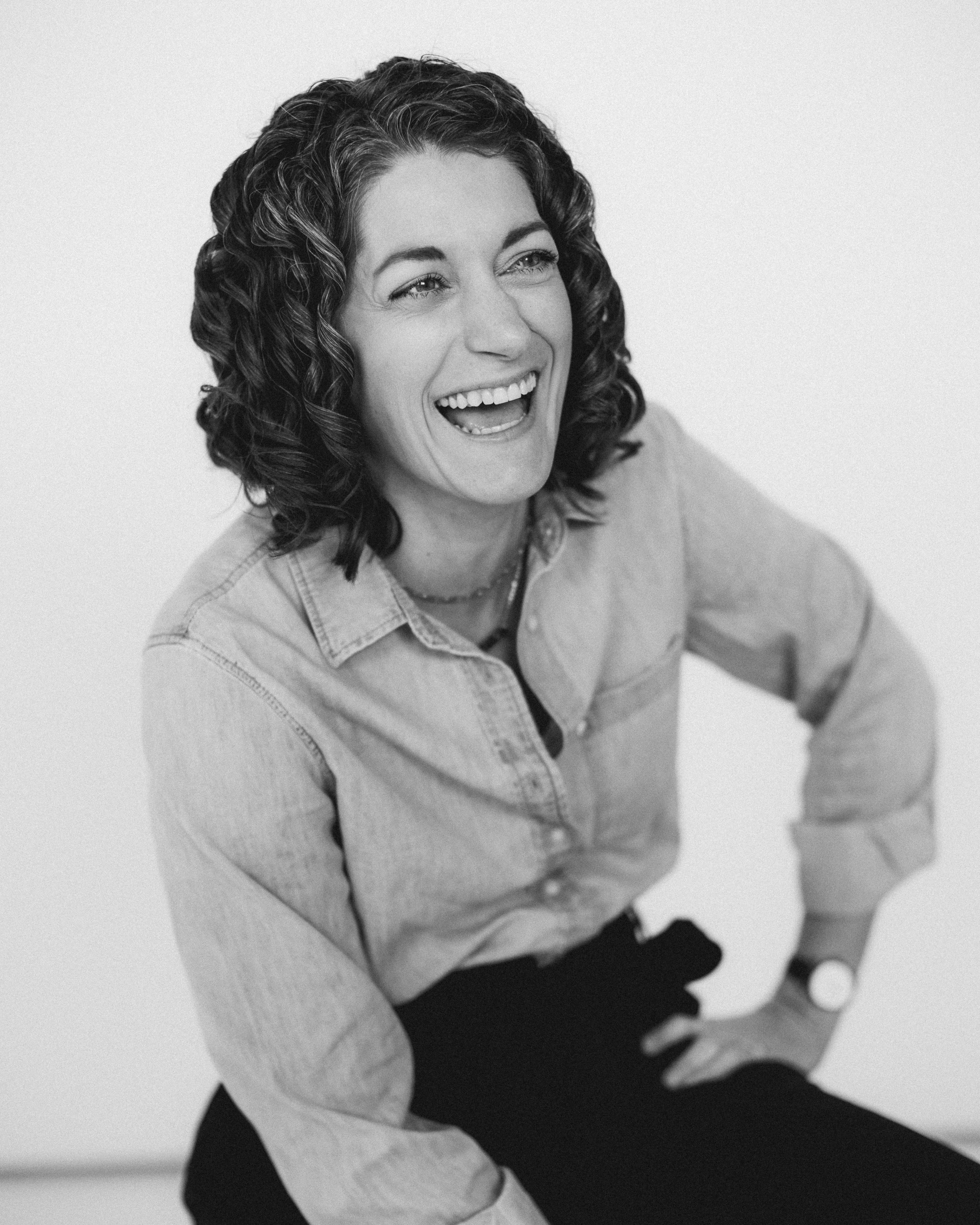 A woman with curly hair laughing, wearing a denim shirt and black pants.