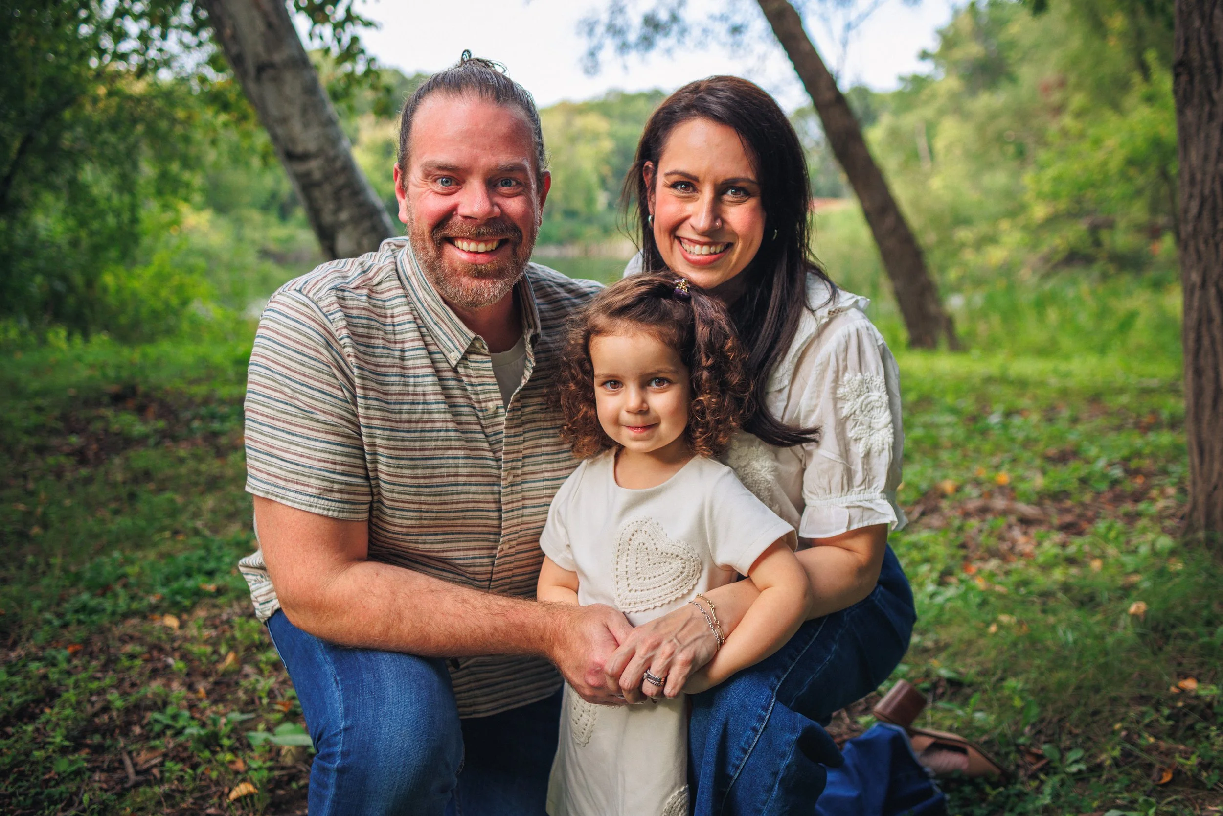 A happy family of three, a man, a woman, and a young girl, smiling and posing outdoors in a wooded area with green trees and grass background.