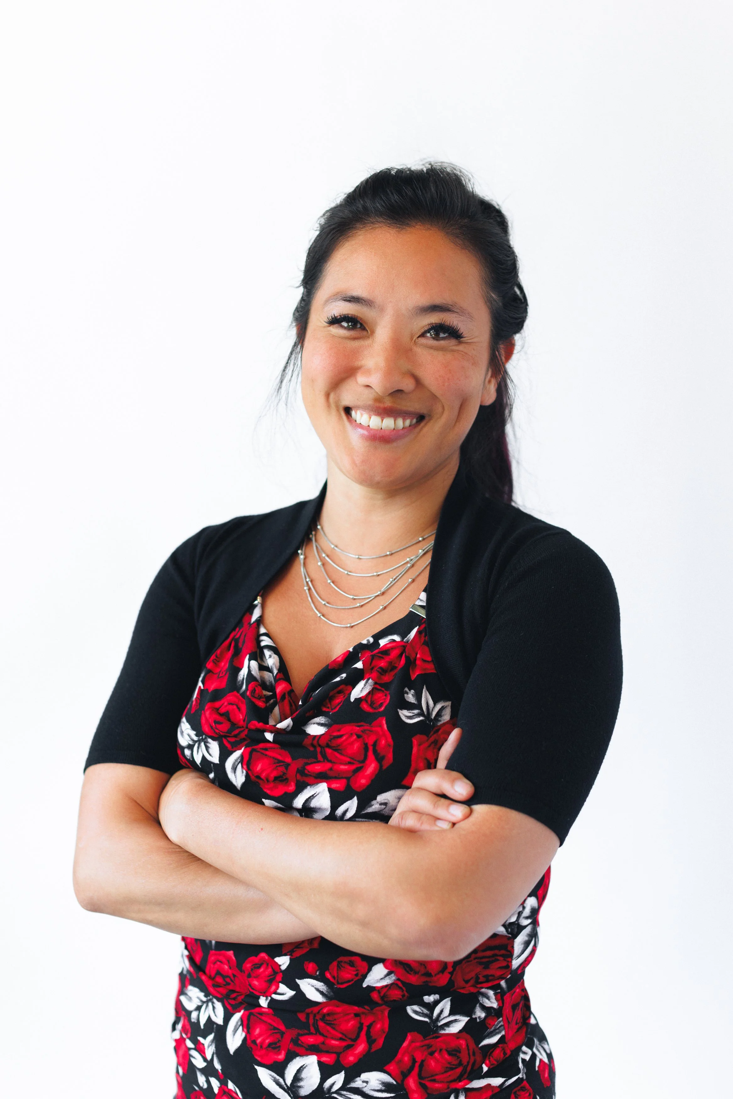 A woman with long dark hair smiling and crossing her arms, wearing a black cardigan over a red and black floral dress, and layered silver necklaces, standing against a white background.