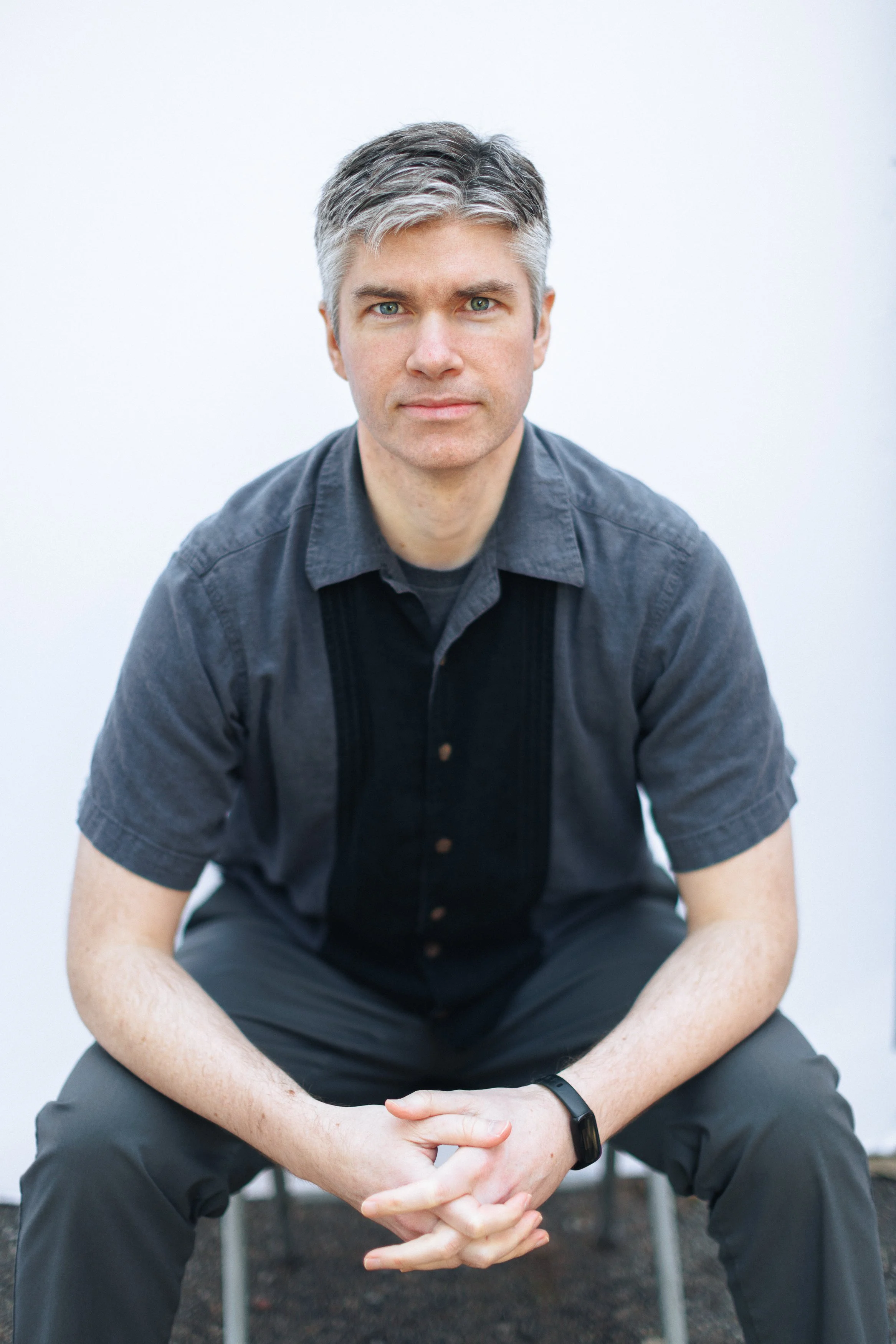 A man with short, gray hair and fair skin sitting on a stool against a plain white background. He wears a short-sleeved dark shirt and dark pants, with a black watch on his left wrist. He is looking directly at the camera with a neutral expression.