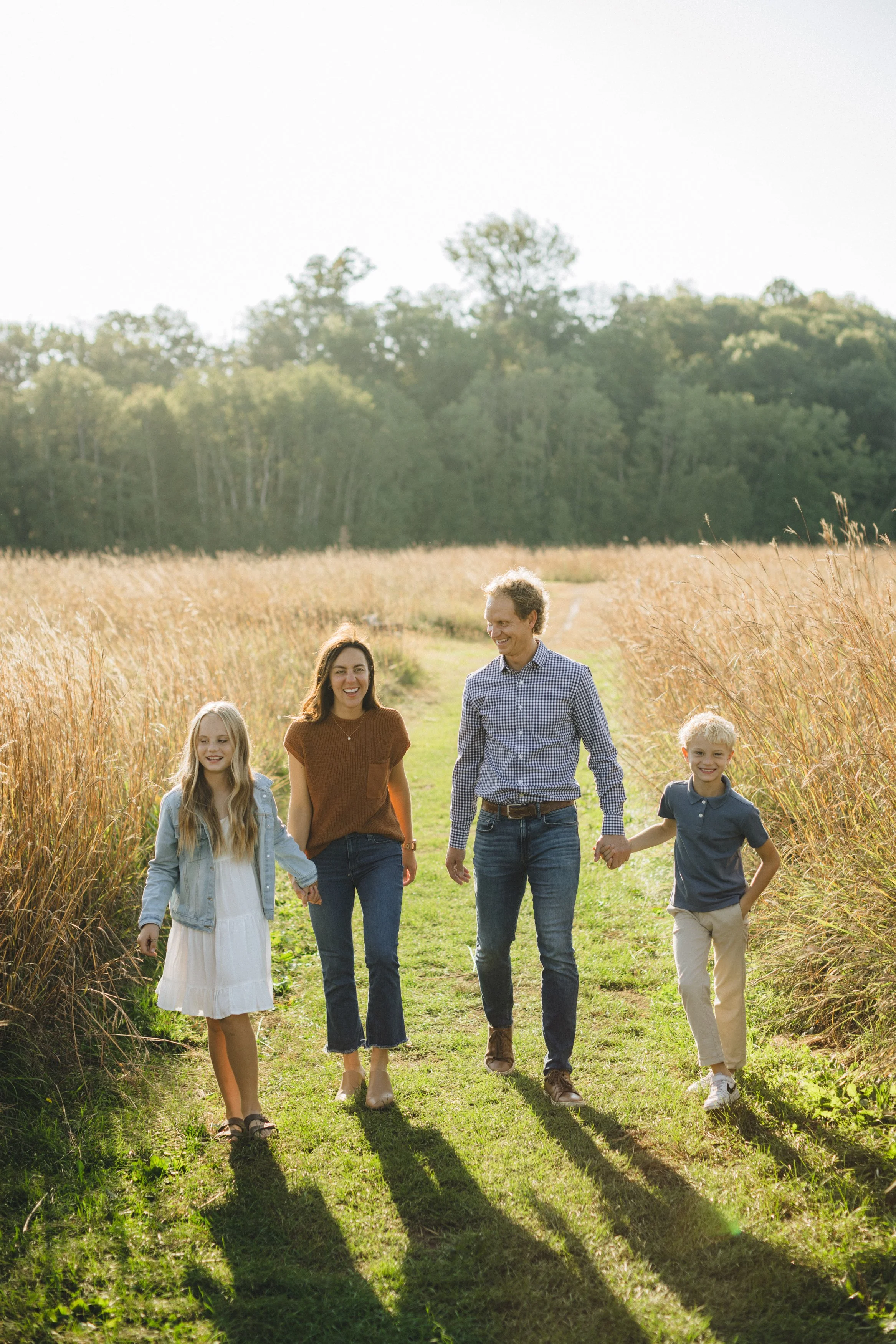 A family of four walking hand-in-hand in a grassy field with tall golden grasses and trees in the background on a sunny day.