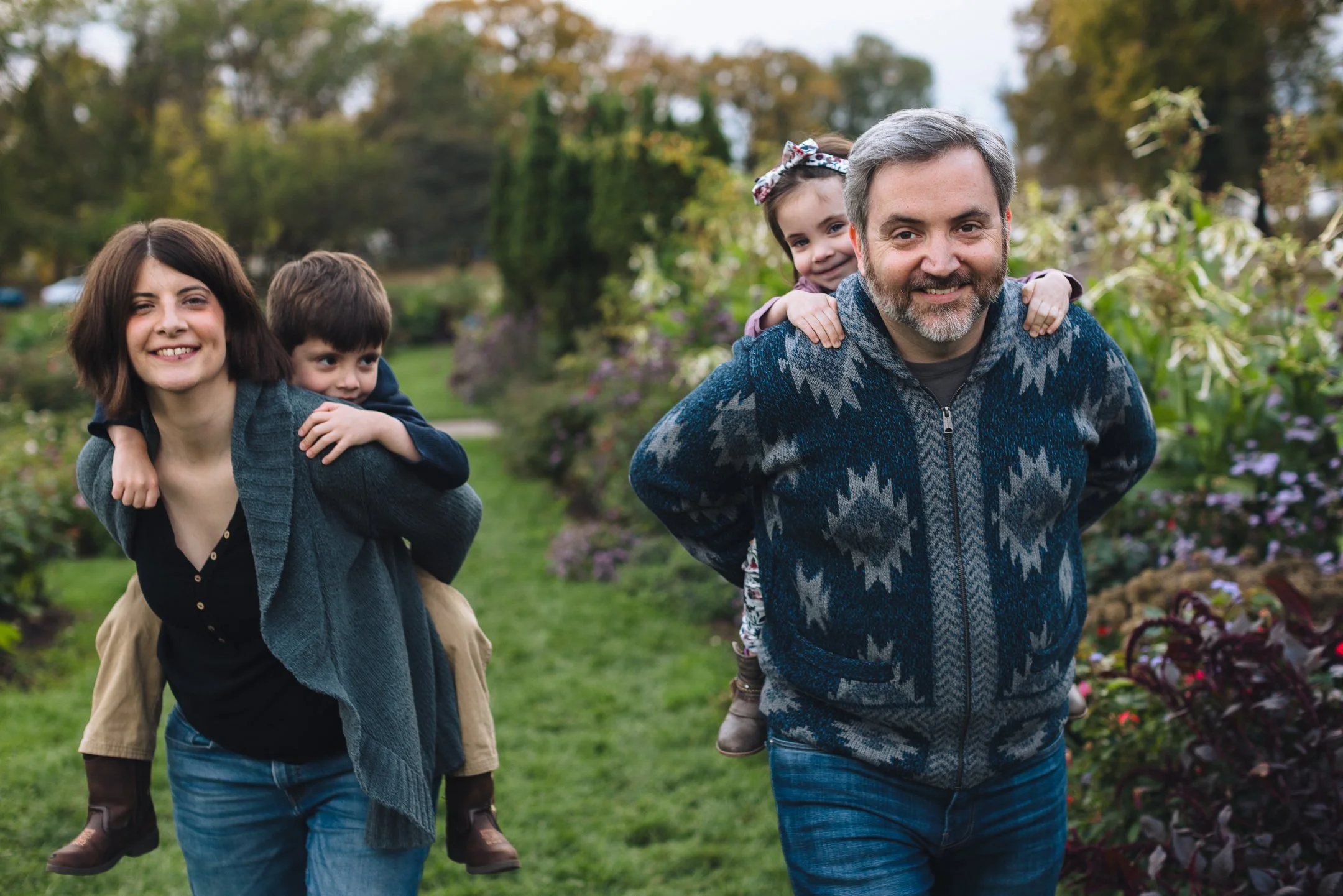 A family of four with two adults and two children walking outdoors in a park or garden, smiling and enjoying a sunny day.