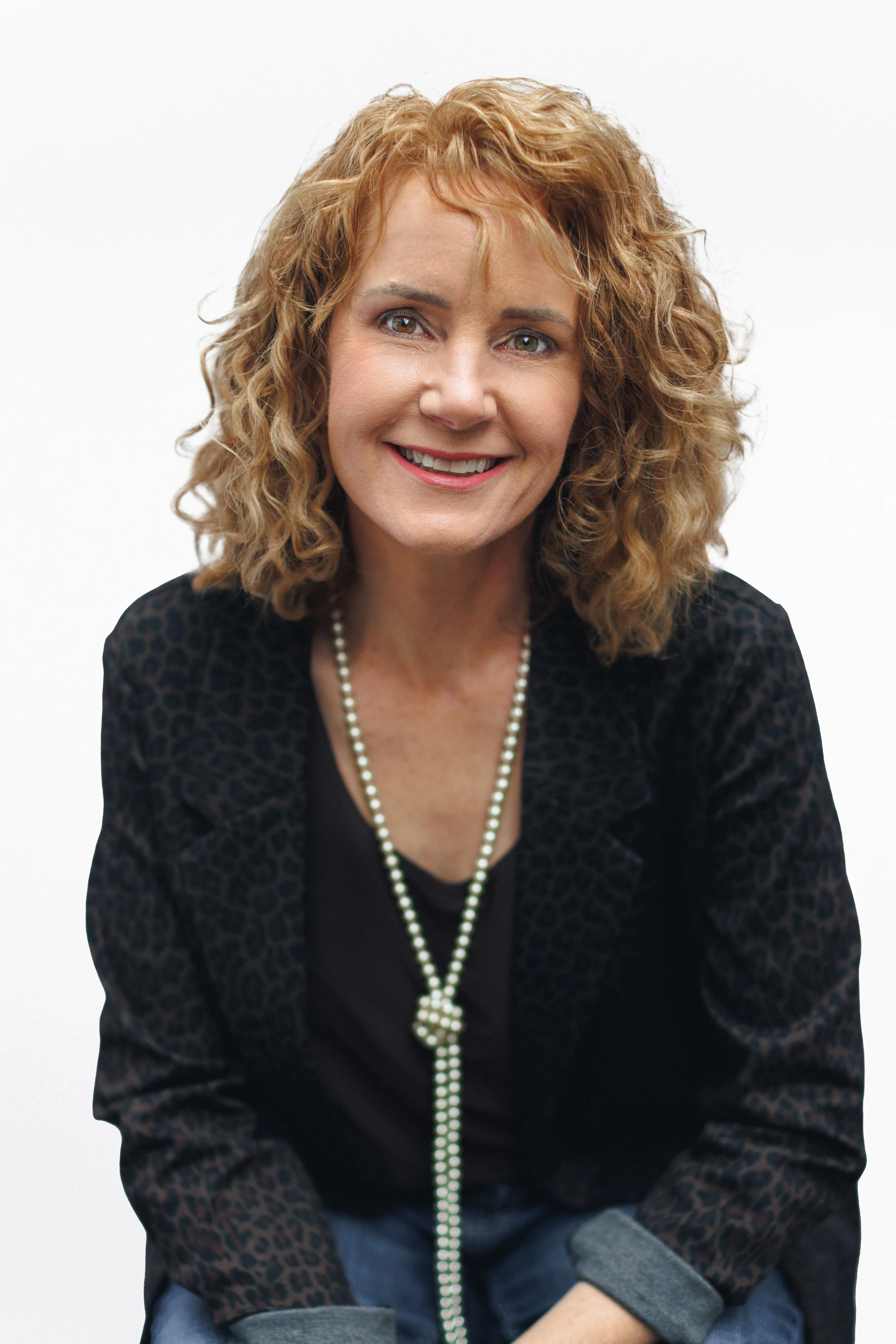 A woman with curly red hair smiling, wearing a black top, animal print blazer, and a long pearl necklace, sitting against a plain white background.