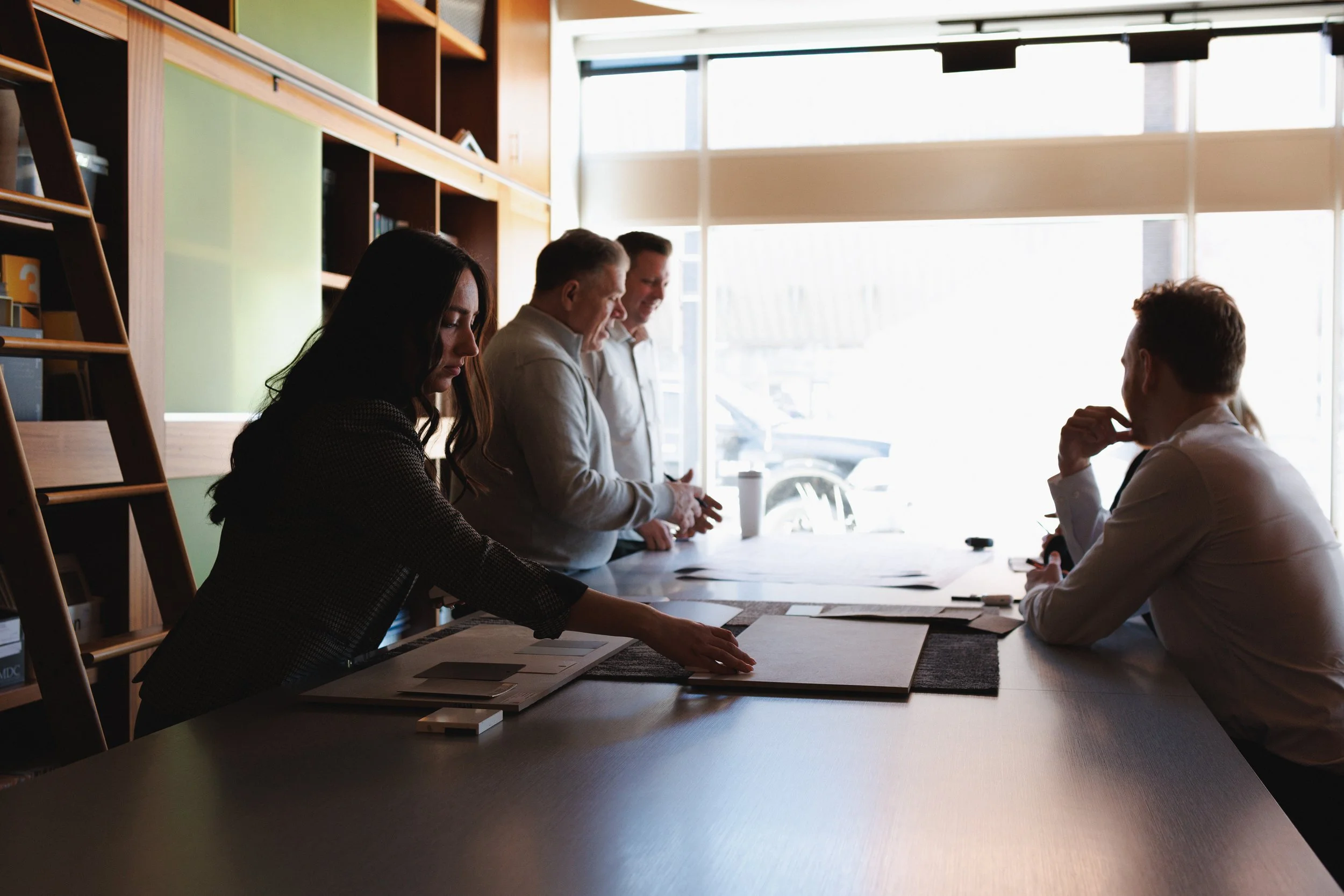 A group of five professionals in a business meeting or discussion around a conference table in a bright, modern office with large windows, bookshelves, and natural light.
