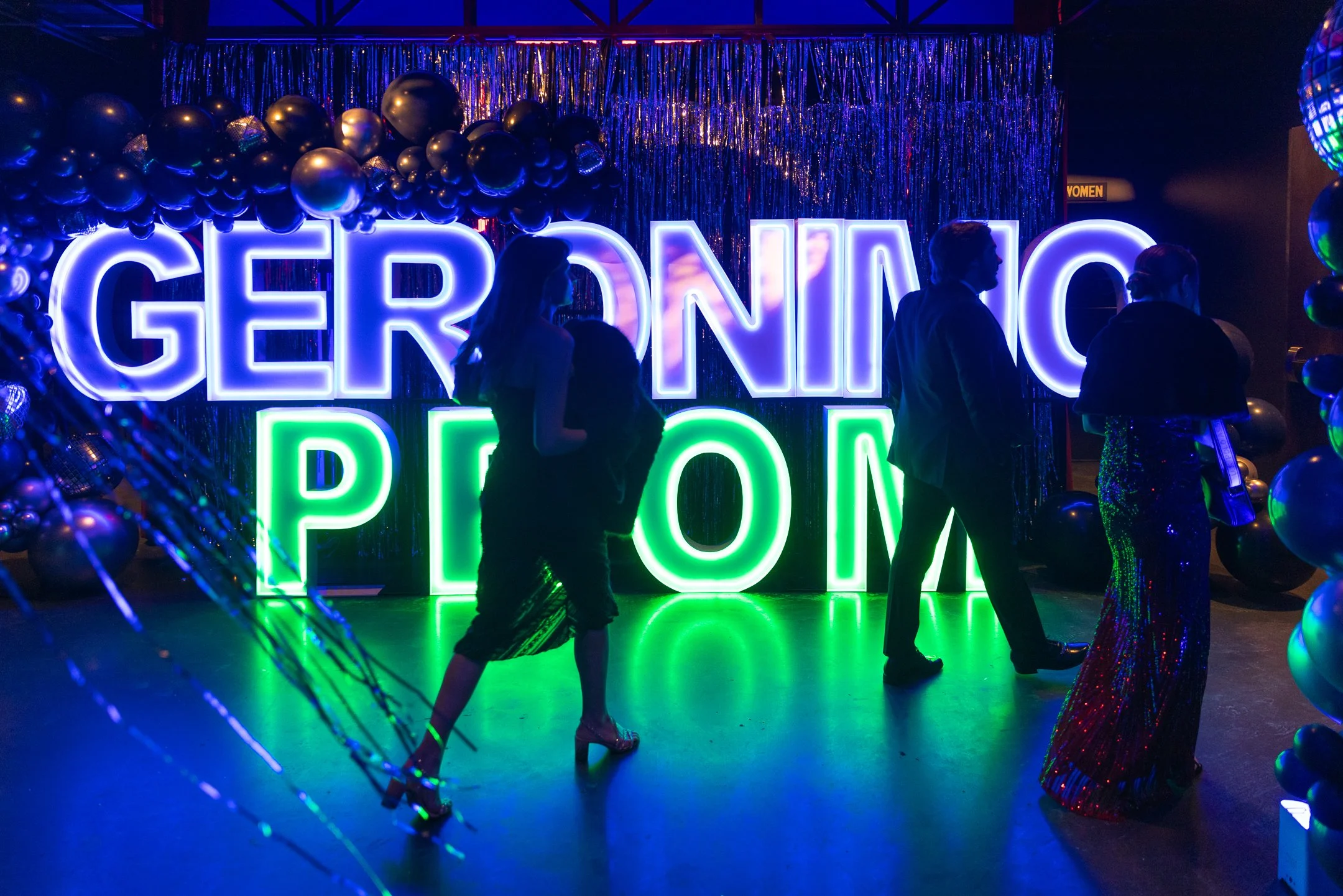 Silhouettes of people at a neon-lit party with the words 'GERRONIMO POP' in bright, colorful lights, surrounded by balloons and metallic decorations.