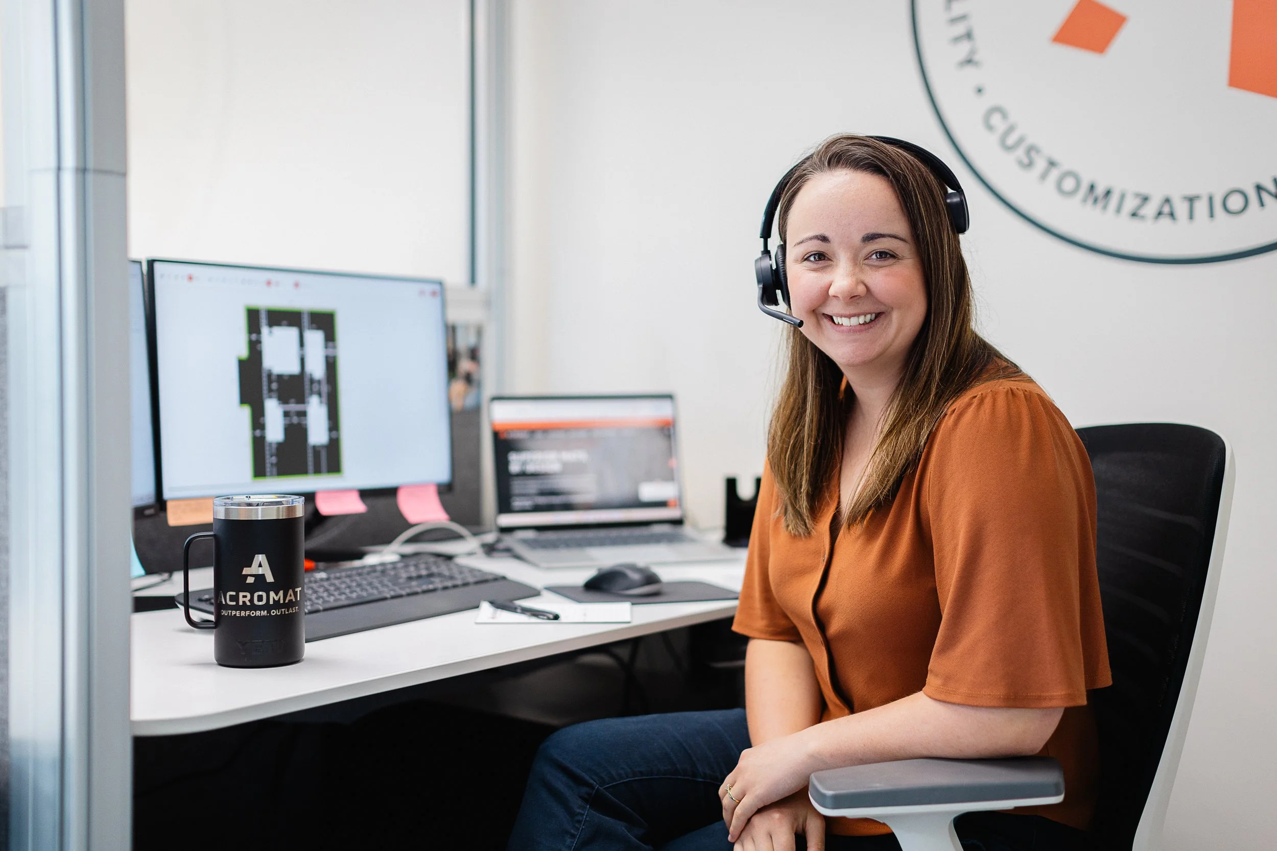 A smiling woman with brown hair wearing a headset, sitting at a desk with multiple computer screens in an office. The desk has a black travel mug and a computer keyboard.