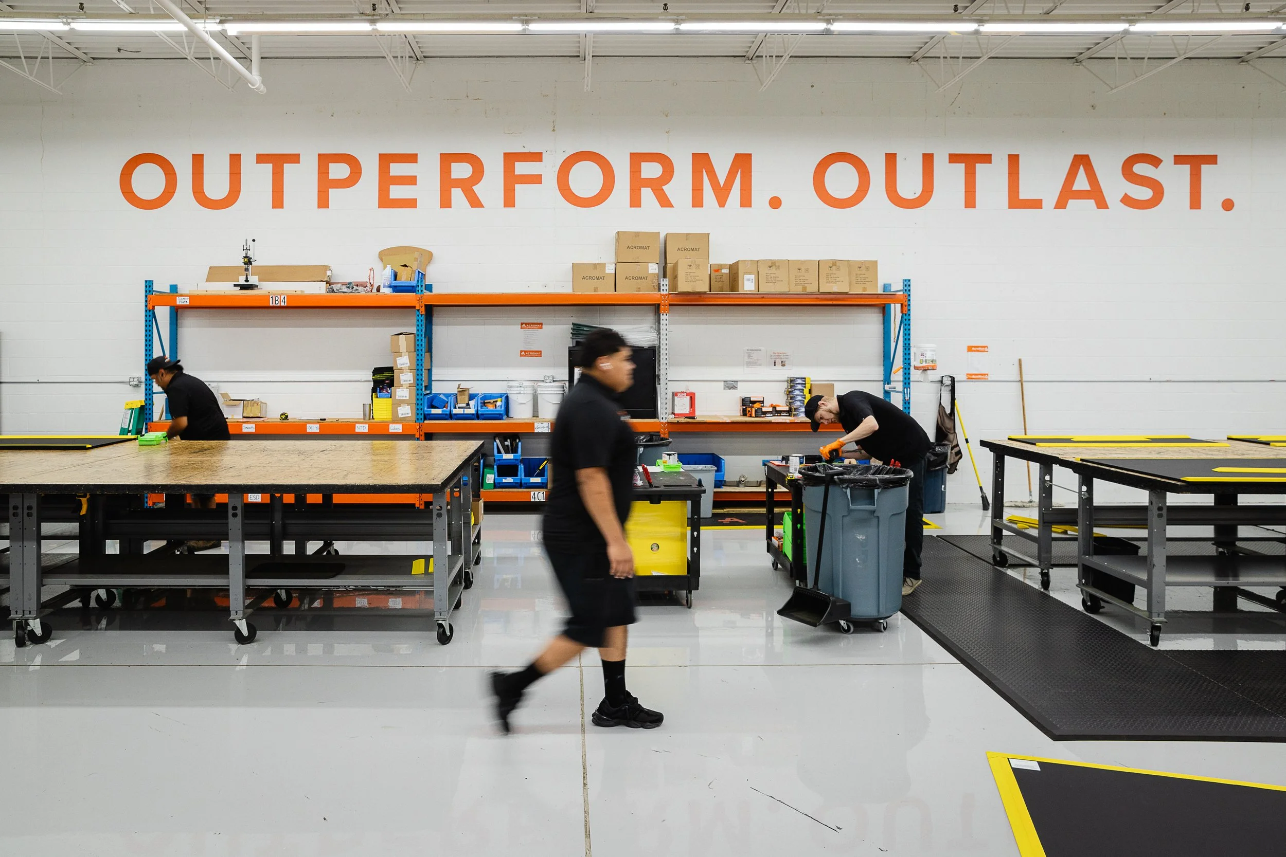 Inside a warehouse or manufacturing facility with large orange and blue shelving units, workers organizing or assembling items, and a wall with a large orange sign that reads 'OUTPERFORM. OUTLAST.'