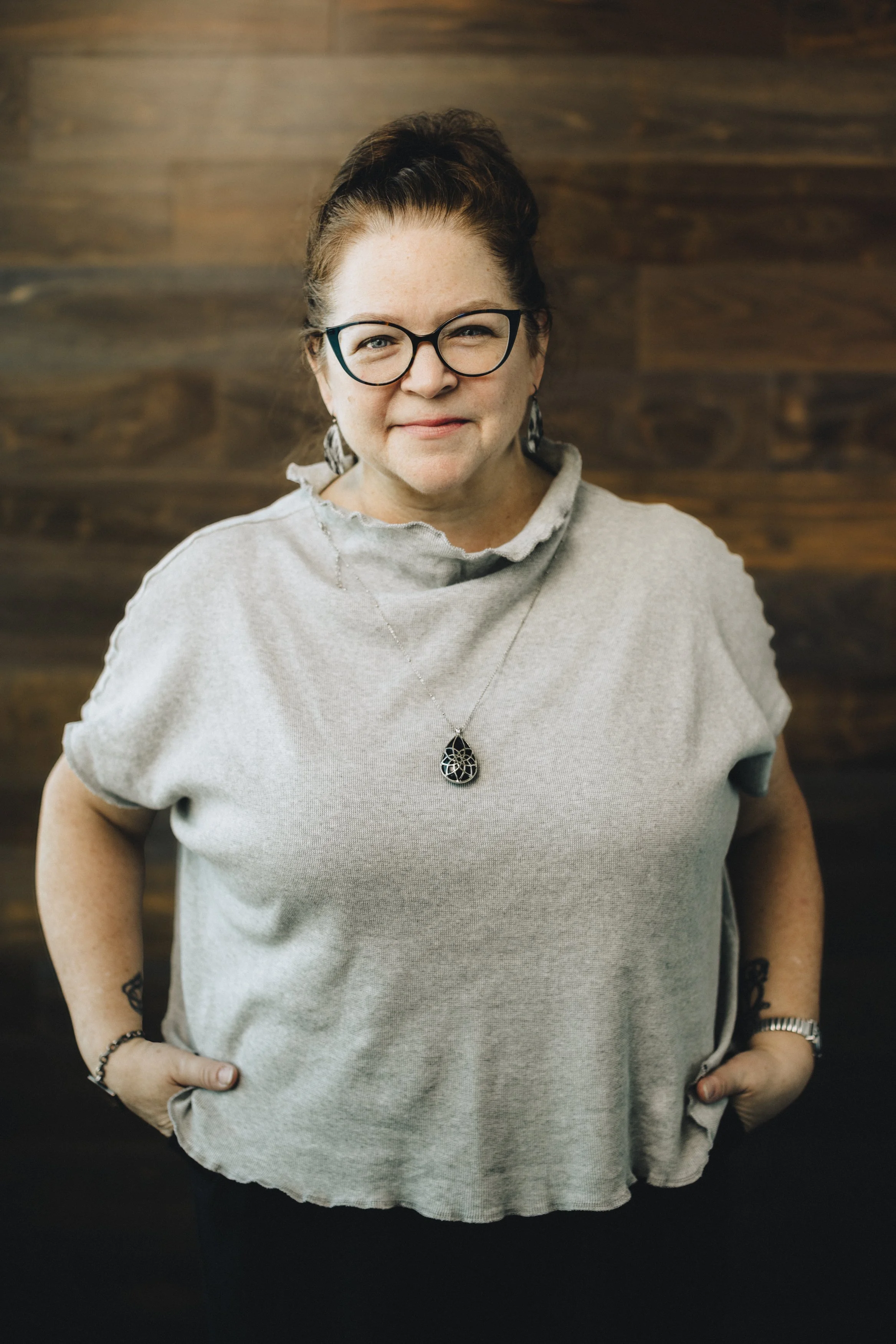 A woman with glasses and red hair tied back, wearing a light gray, short-sleeved top with frayed collar, standing with hands in pockets against a wooden-paneled wall.