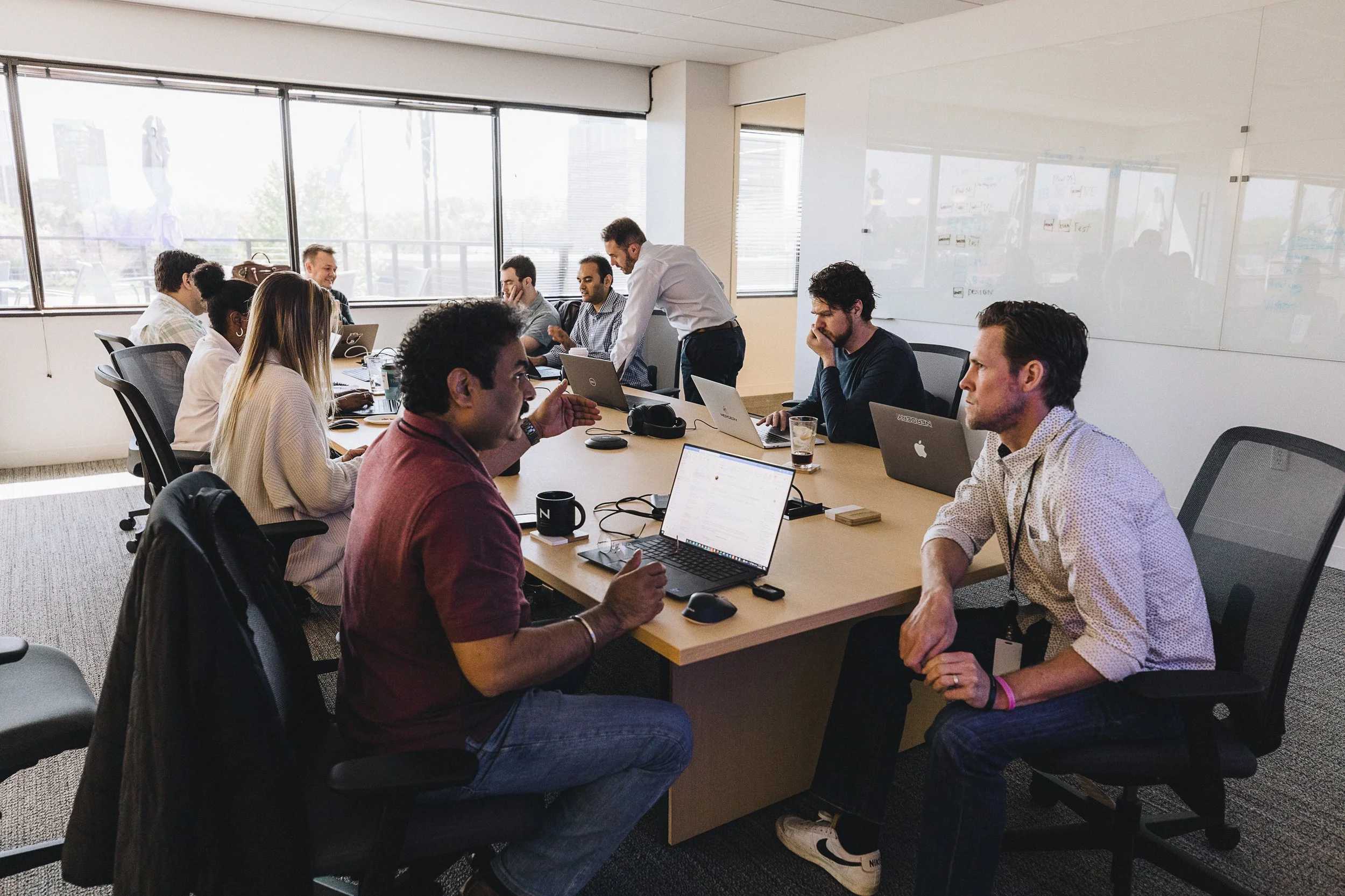 Business professionals in a meeting room, seated around a large table with laptops, discussing. One person standing and talking, others listening attentively.