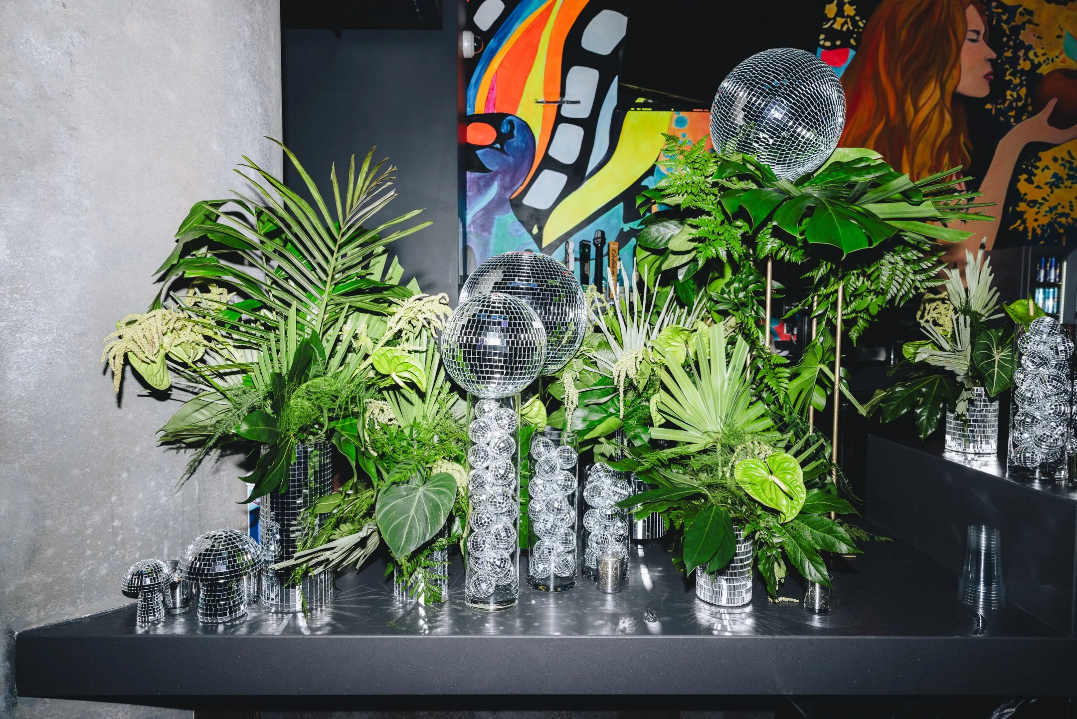 Arrangement of green tropical plants in mirrored vases, with disco balls on sticks, on a black table in front of colorful urban art murals.
