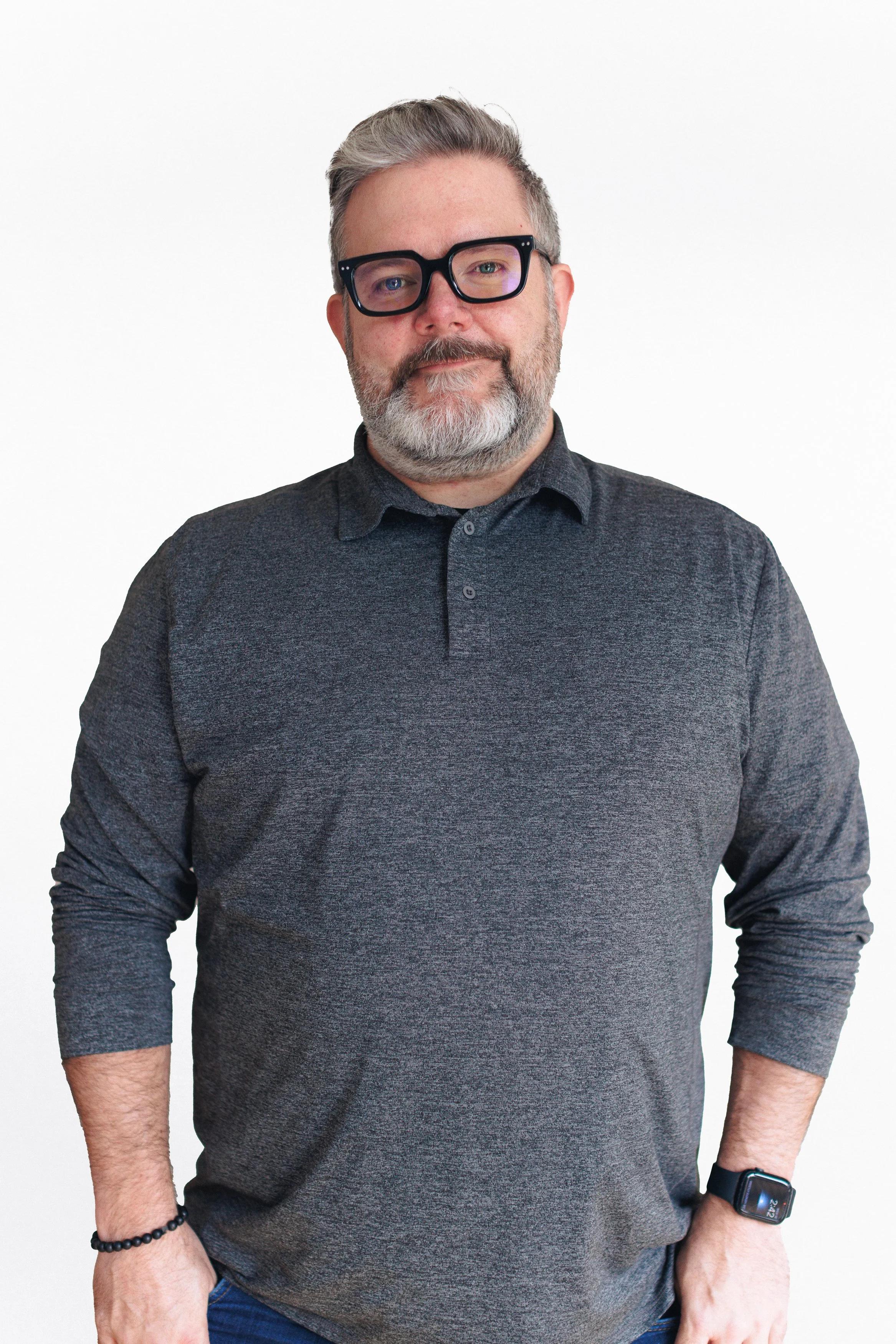 Man with gray hair and beard wearing black glasses, black shirt, and smartwatch posing against a white background.