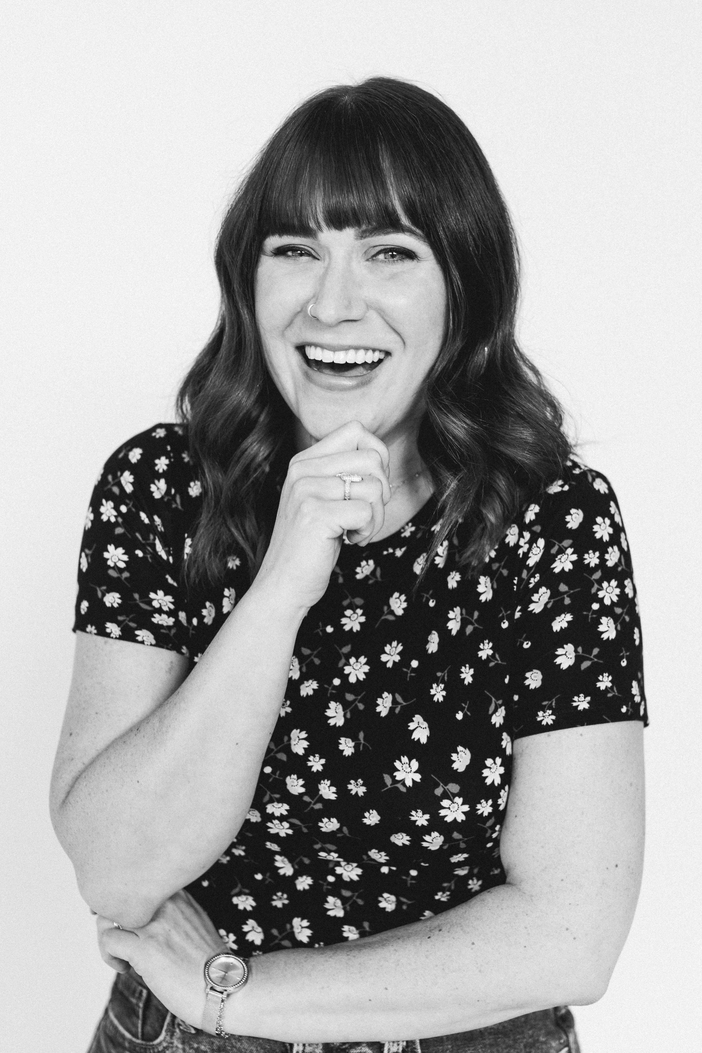 A smiling woman with shoulder-length wavy hair and a nose ring, wearing a floral shirt, with her hand near her chin, against a plain background.