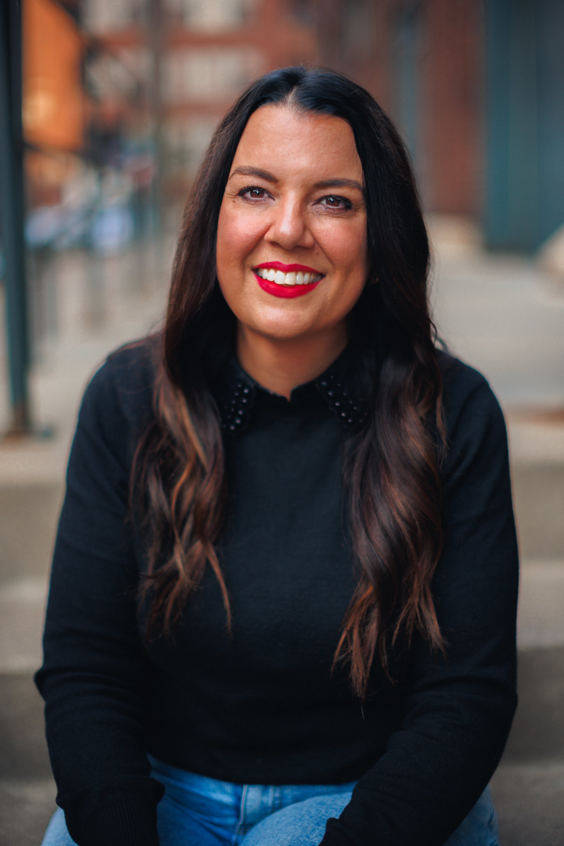 A woman with long dark wavy hair, red lipstick, and a black top with polka dot collar, smiling outdoors.