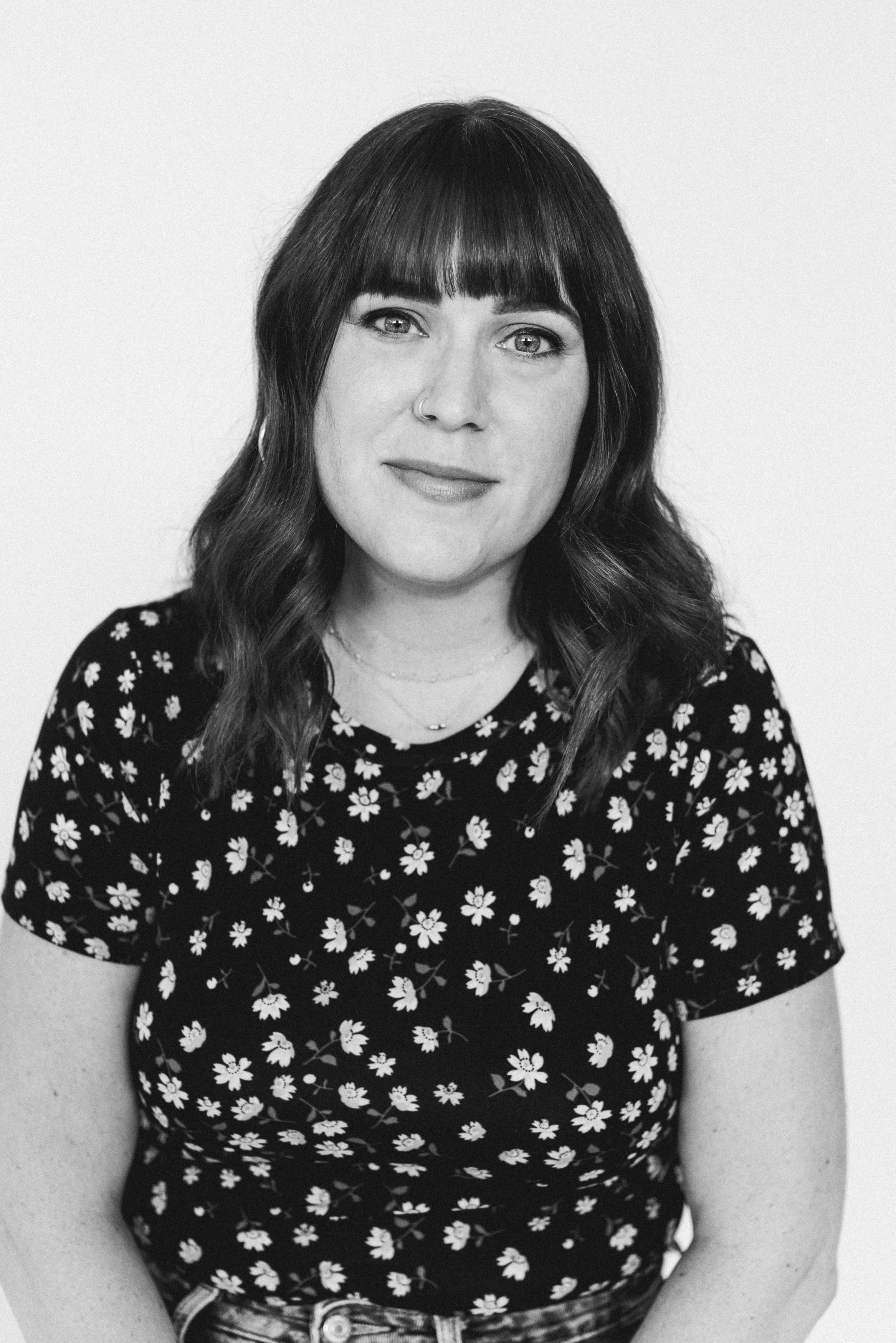 Black and white portrait of a woman with shoulder-length wavy hair and blunt bangs, wearing a floral patterned shirt and smiling slightly, against a plain light background.