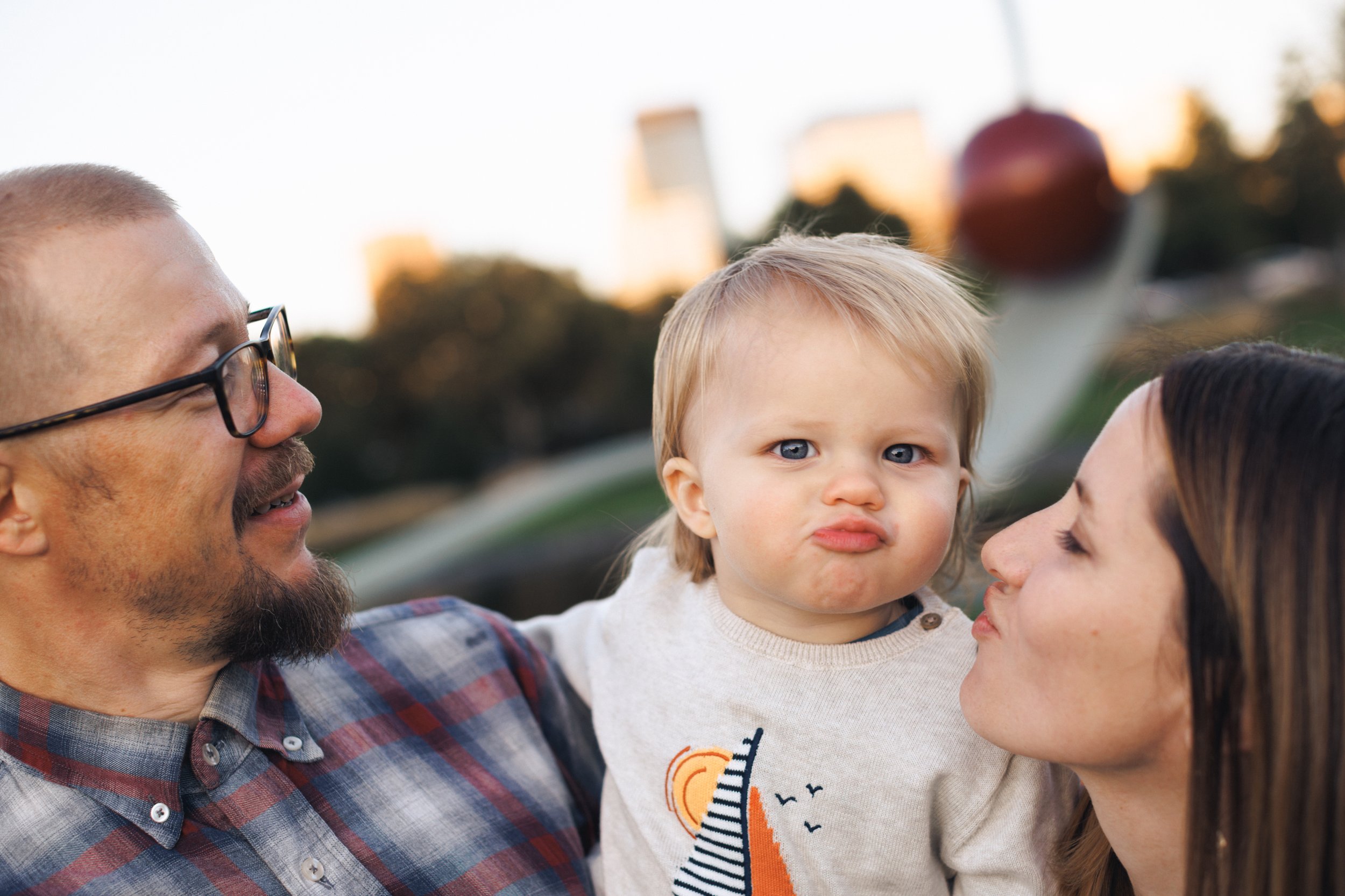 A family outdoor portrait with a man, woman, and young child, with the child making a pouty face, during the evening with city buildings in the background.