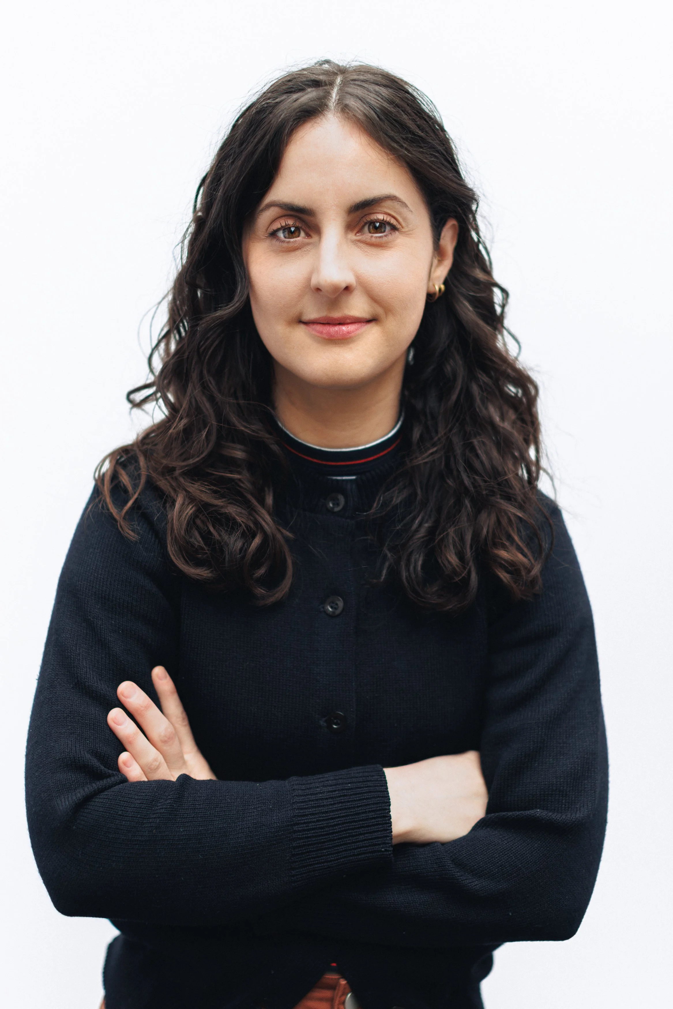 A woman with wavy dark hair, crossed arms, and a confident expression, standing against a plain white background.