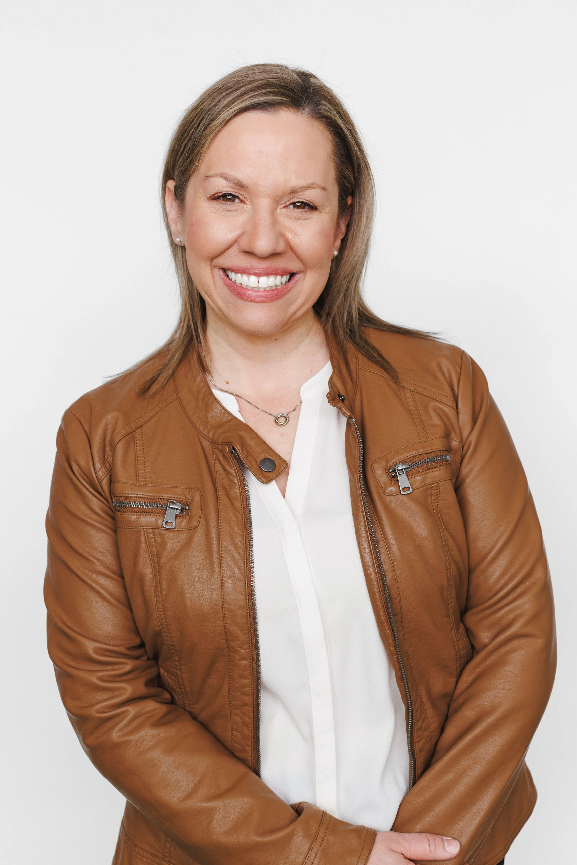 A woman with shoulder-length blonde hair, wearing a brown leather jacket over a white blouse, smiling at the camera against a plain white background.