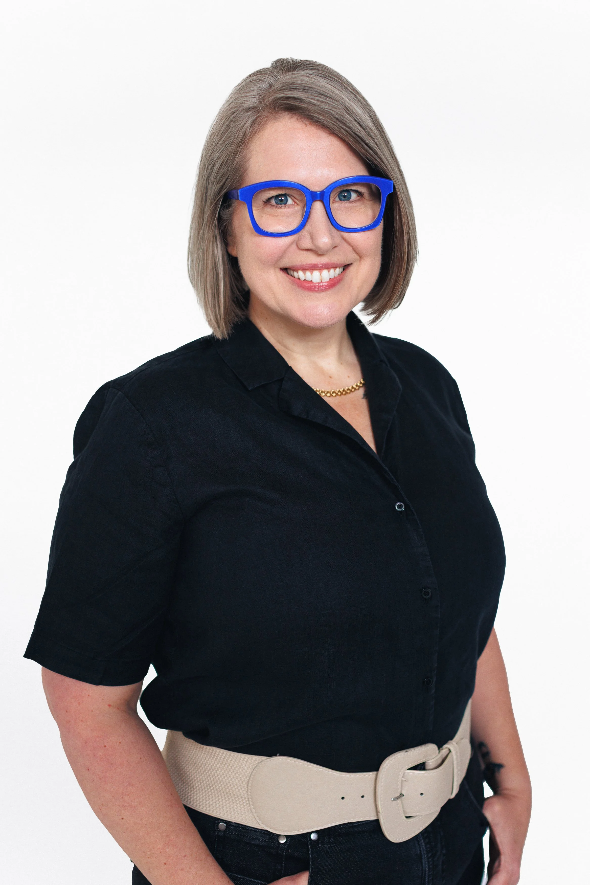 A woman with shoulder-length gray hair, wearing blue glasses, a black button-up shirt, a gold necklace, and a beige belt, smiling against a white background.