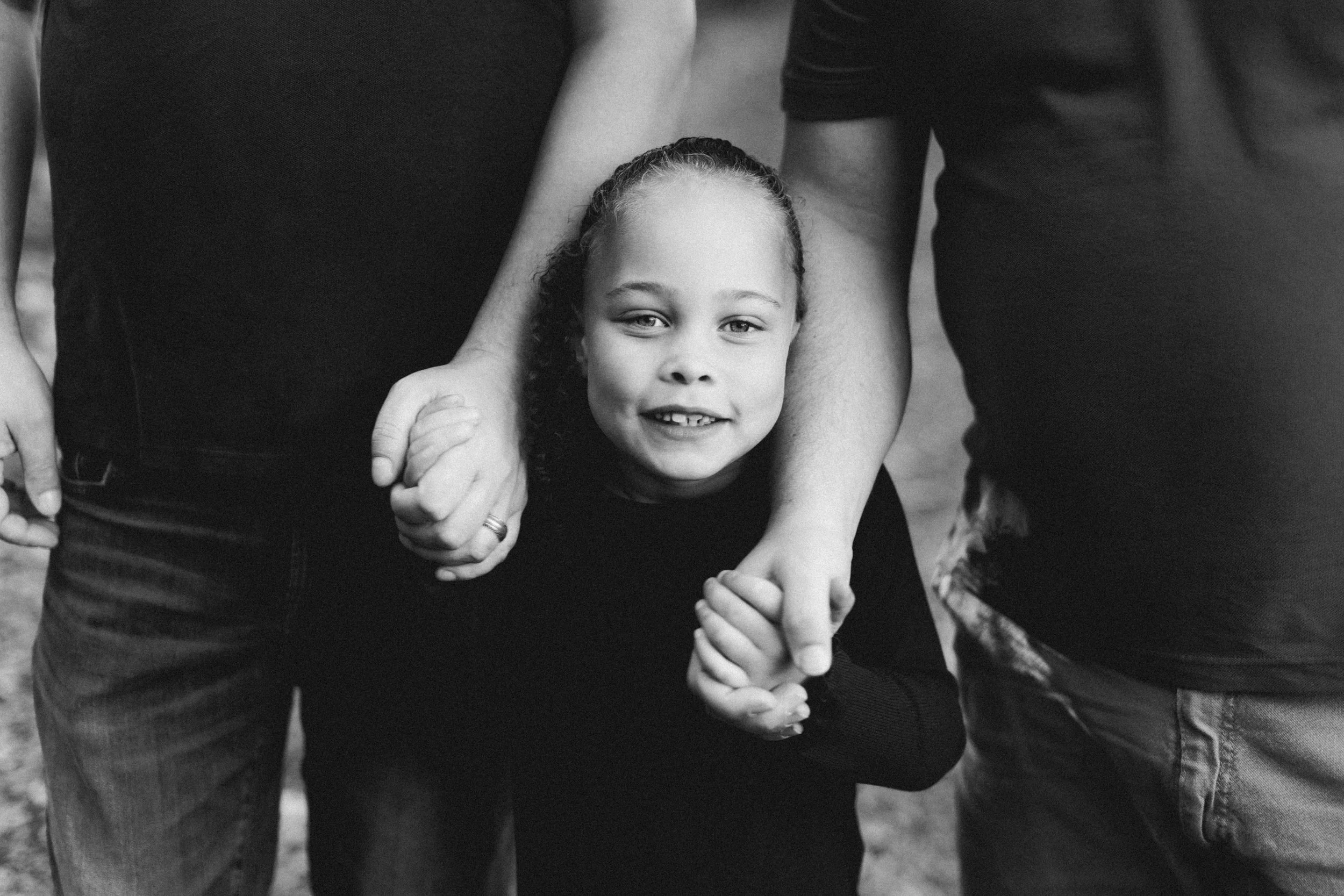 A young girl with curly hair smiling, holding hands with two adults, standing outdoors.