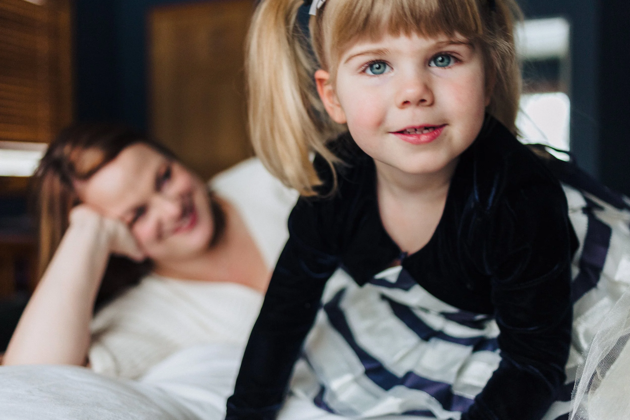 A young girl with blue eyes and blond hair looks at the camera while leaning forward on a bed. A woman, likely her mother, is in the background smiling and lying on the bed.