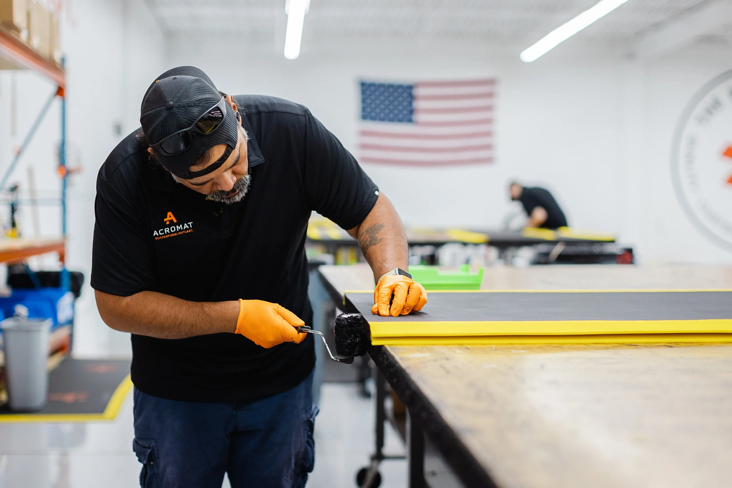 Man wearing black cap, sunglasses, and black shirt painting a yellow surface with a small paint roller in a workshop or warehouse. American flag in background.