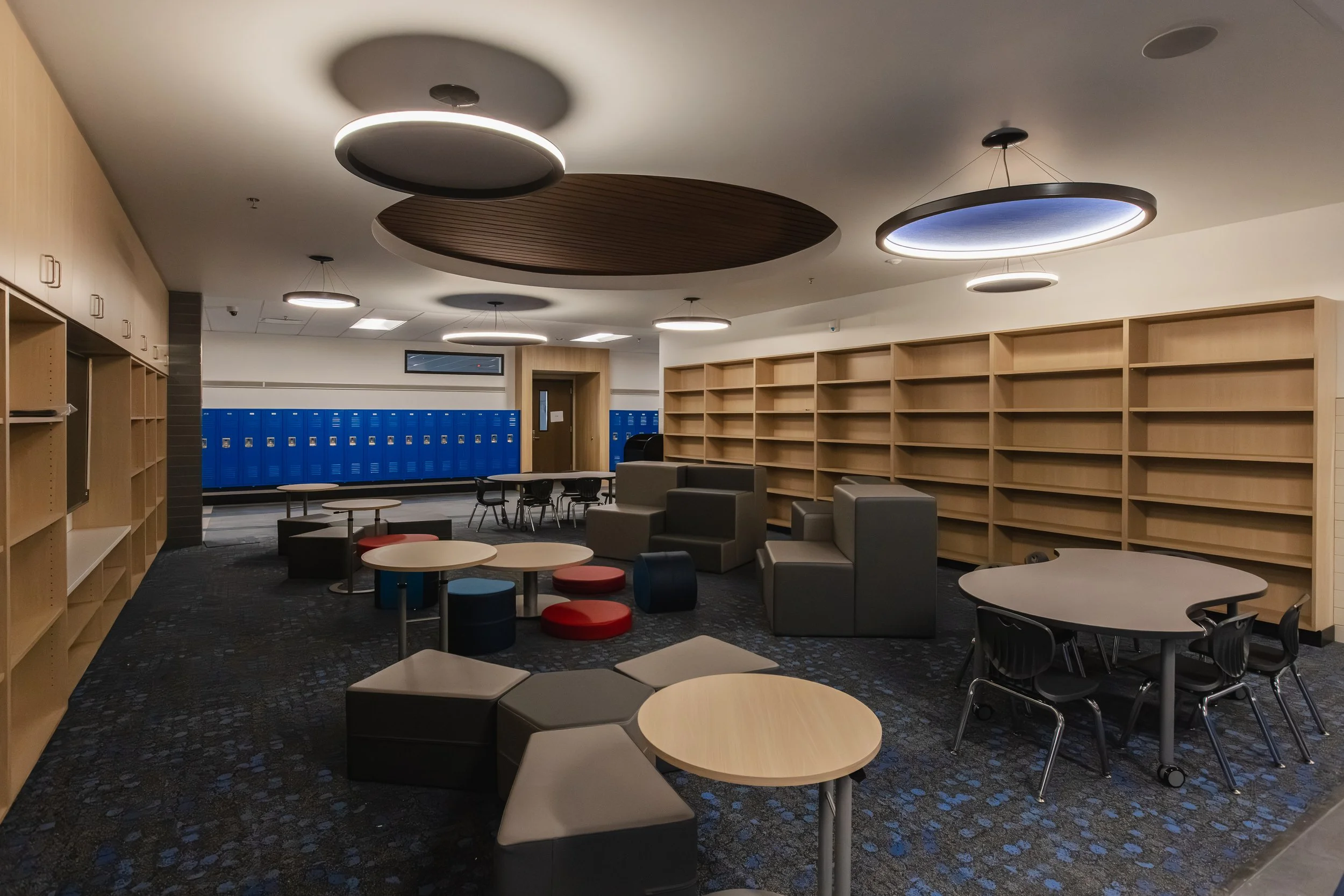 Empty school lounge with blue lockers, wooden bookshelves, tables, and chairs.