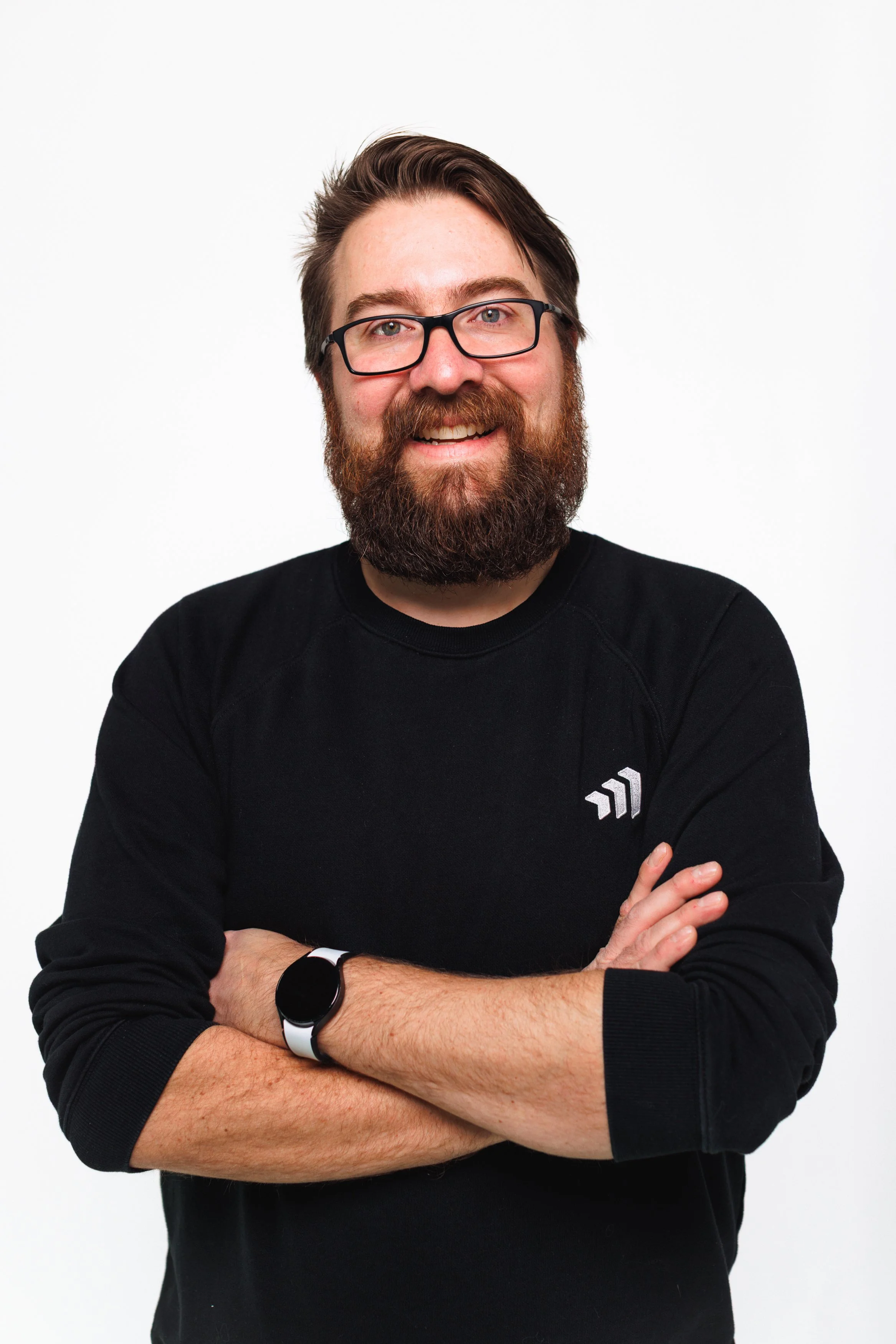 A man with glasses and a beard smiling, wearing a black sweatshirt and a smartwatch, standing against a white background.