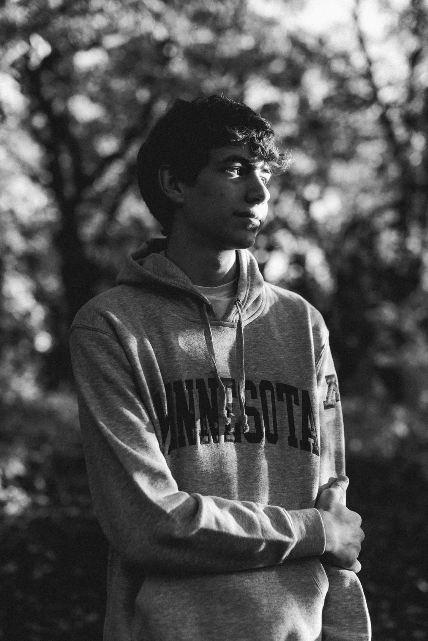 A young man with curly hair wearing a gray hoodie with 'MINNESOTA' printed on it, standing outdoors with blurred trees in the background, in black and white.