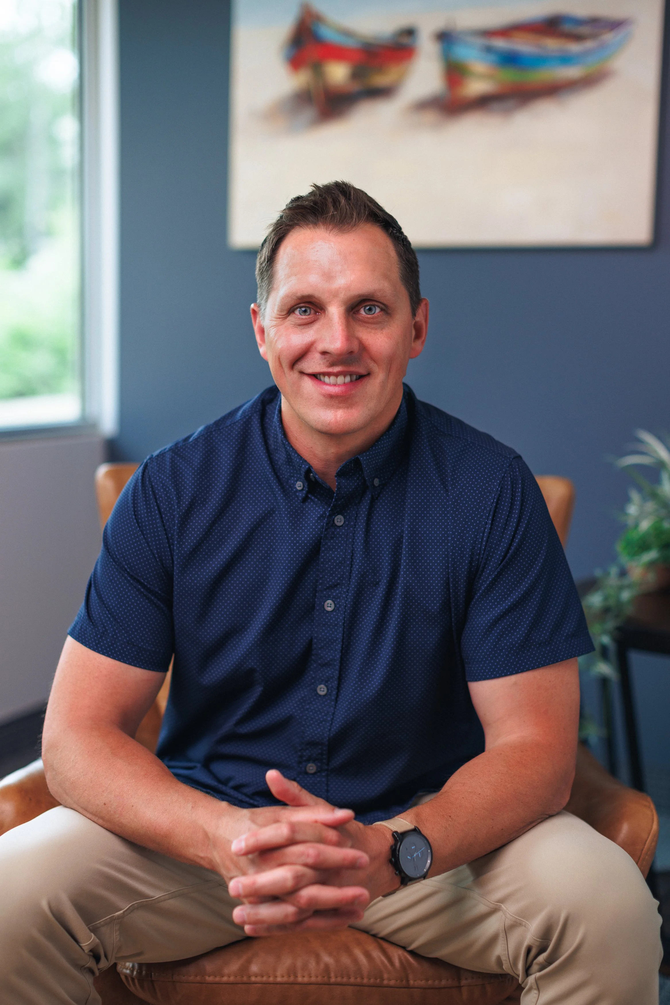 A man sitting on a leather chair in a room with a blue wall, smiling, wearing a blue button-down shirt and a watch, with a window and colorful boat painting in the background.
