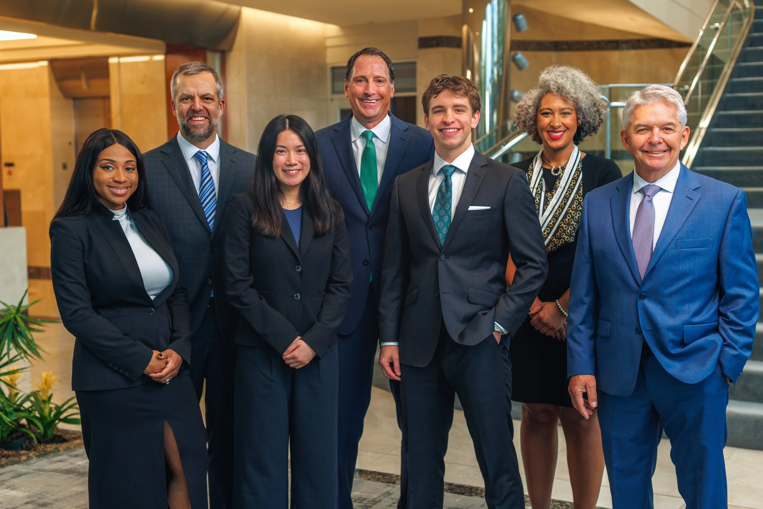 A diverse group of eight professionals in business attire standing together in a modern office lobby, smiling for the camera.
