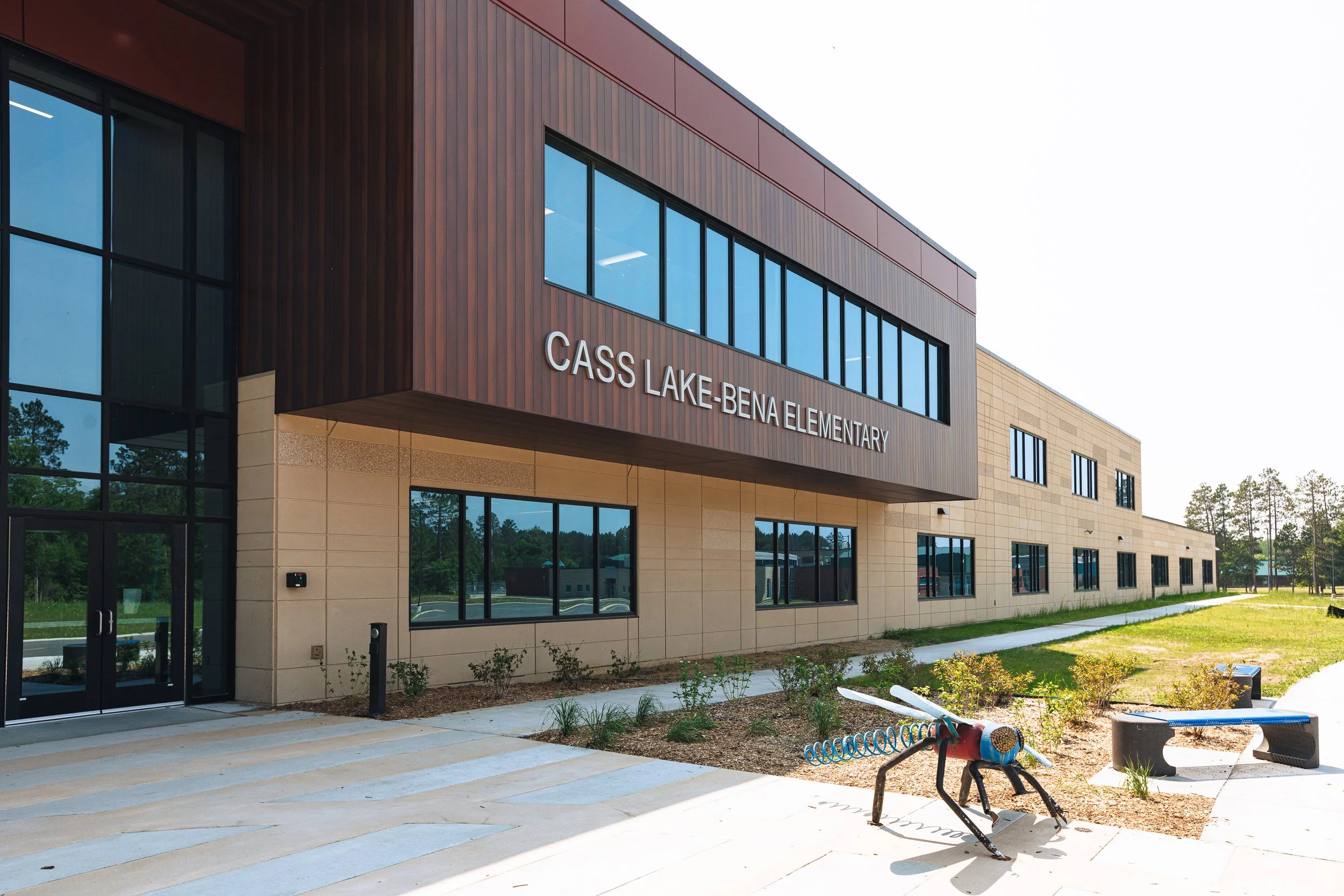Exterior view of Cass Lake-Bena Elementary School with modern architecture, large glass windows, tan and dark brown exterior, and landscaped surroundings, including a colorful metal insect sculpture and benches.