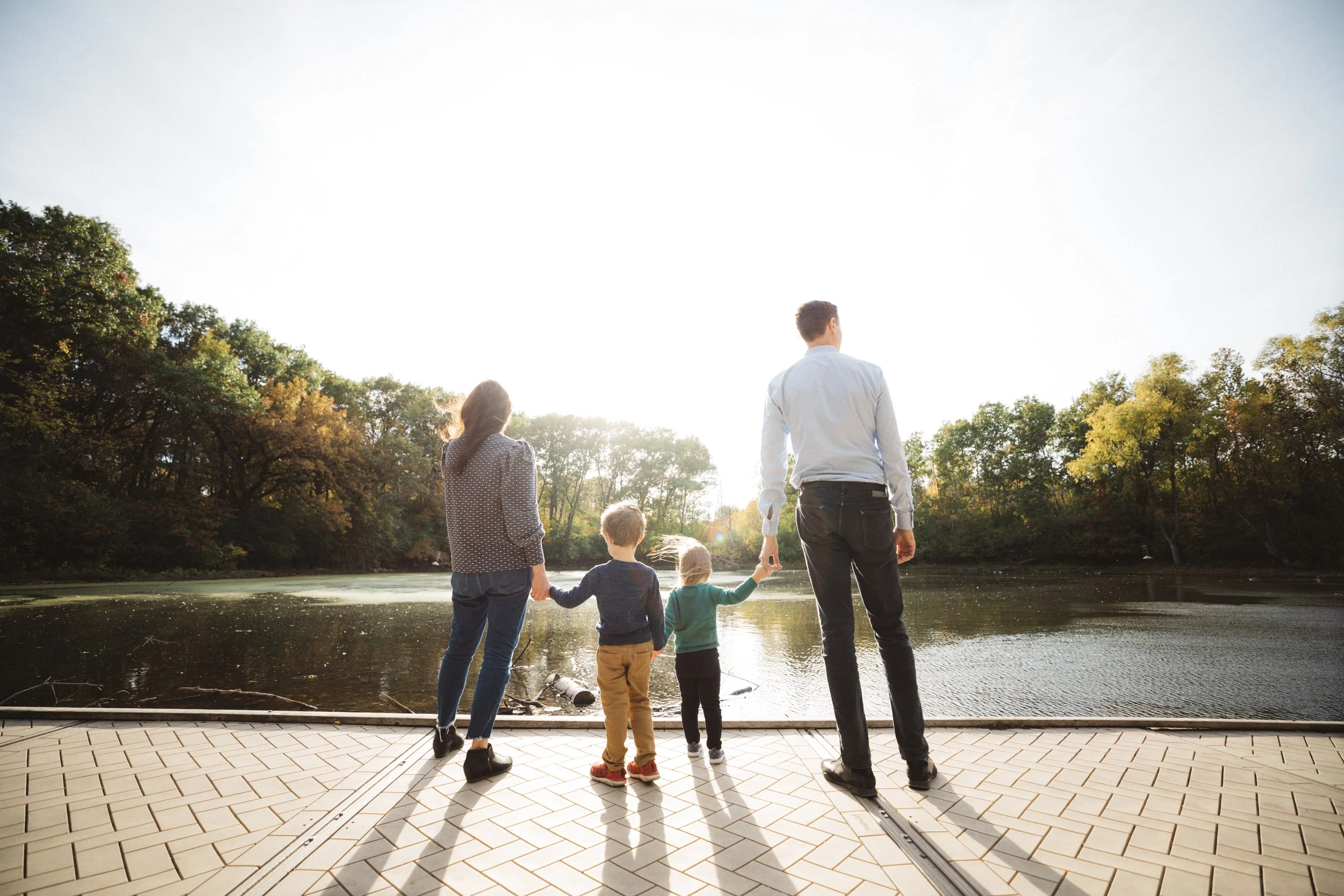 Family of four holding hands and facing a river during sunset