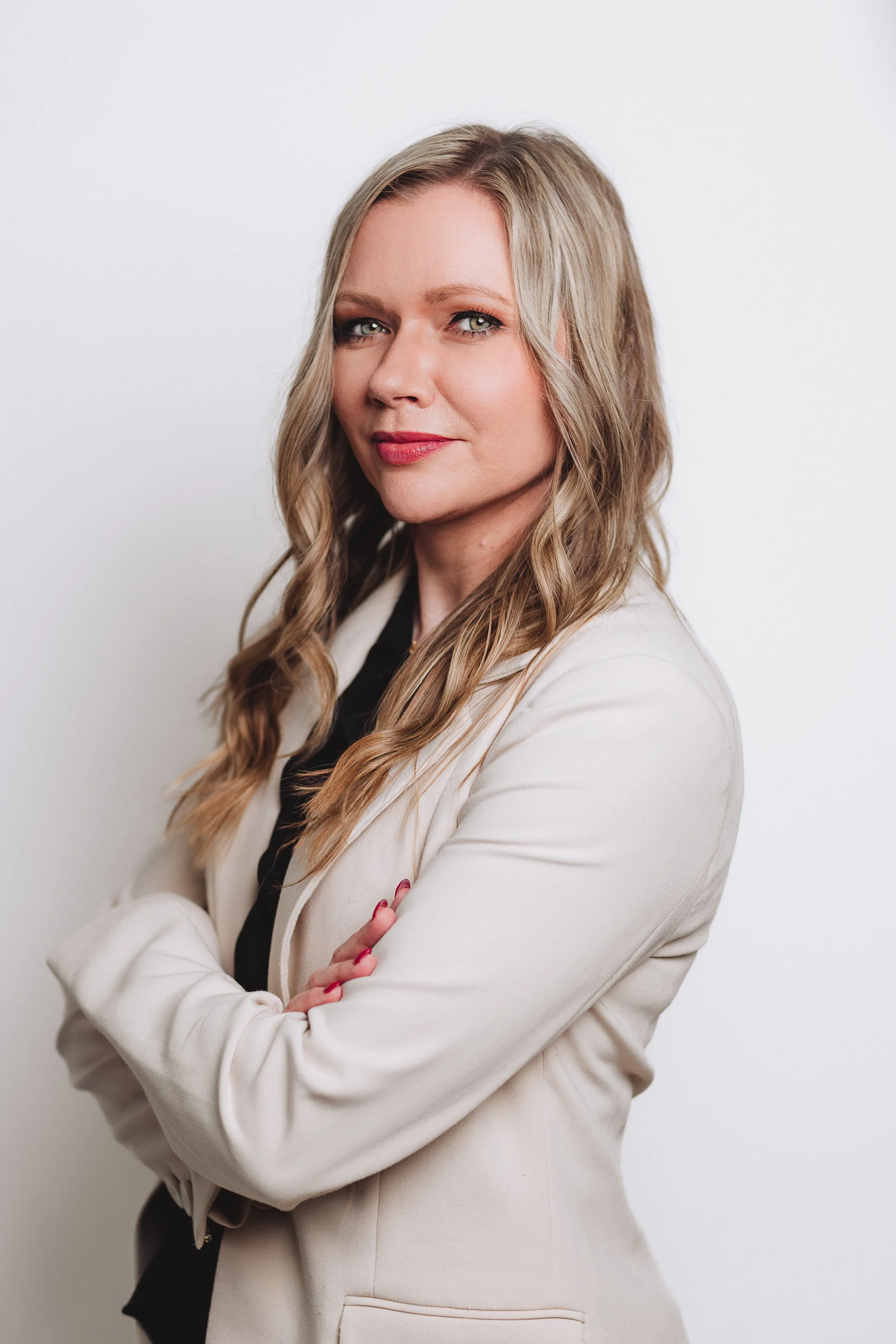 A woman with long, wavy blonde hair and light makeup, wearing a white blazer and black top, standing against a plain white background, with arms crossed and looking confidently at the camera.