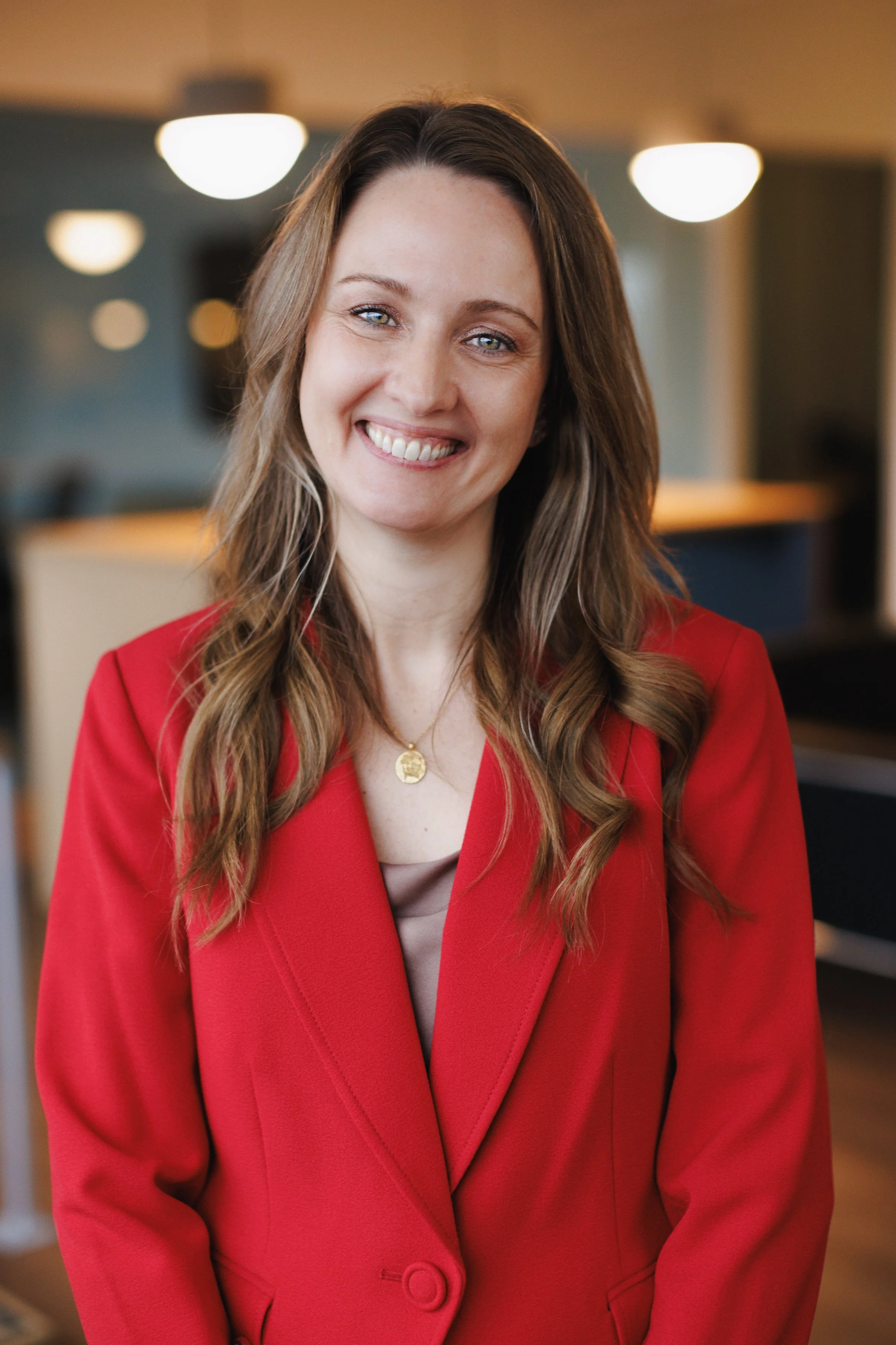 A smiling woman with long, wavy brown hair wearing a red blazer and gold necklace, standing in an indoor setting with warm lighting.