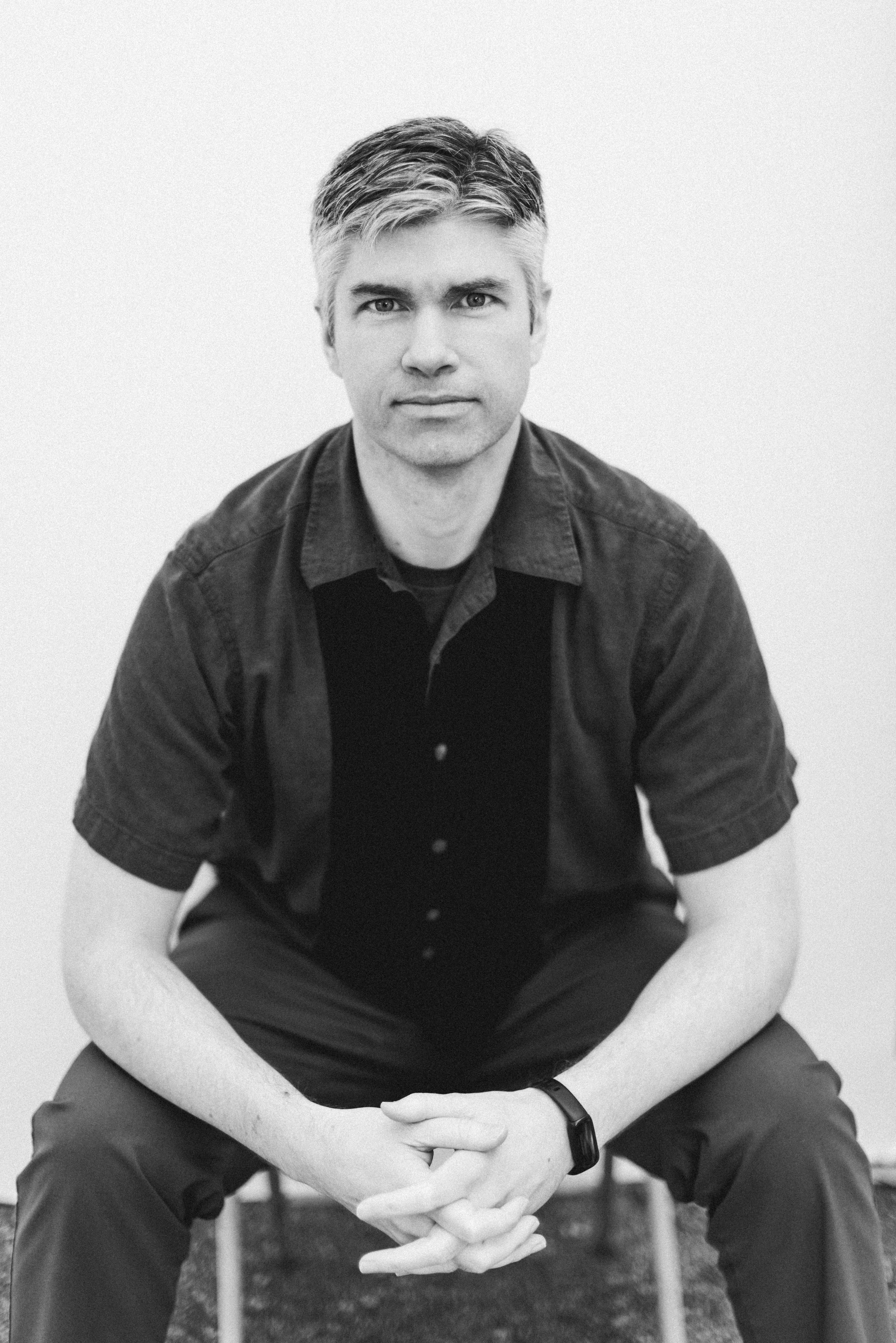 A black-and-white photo of a man sitting with his hands clasped, wearing a short-sleeved button-up shirt and a watch, looking directly at the camera against a plain background.