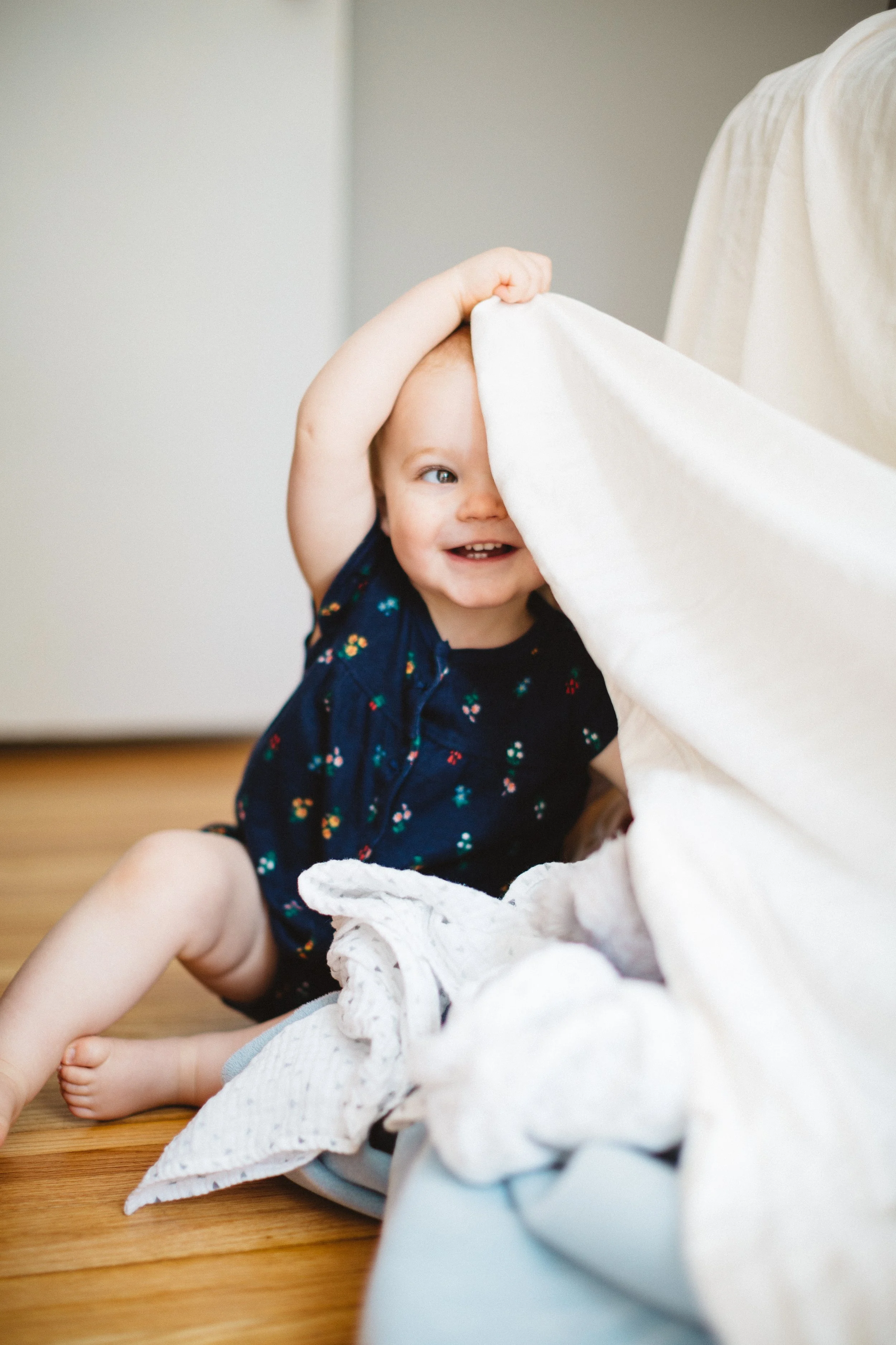 A smiling toddler with a floral patterned navy shirt peeks out from behind a white blanket while sitting on a wooden floor.
