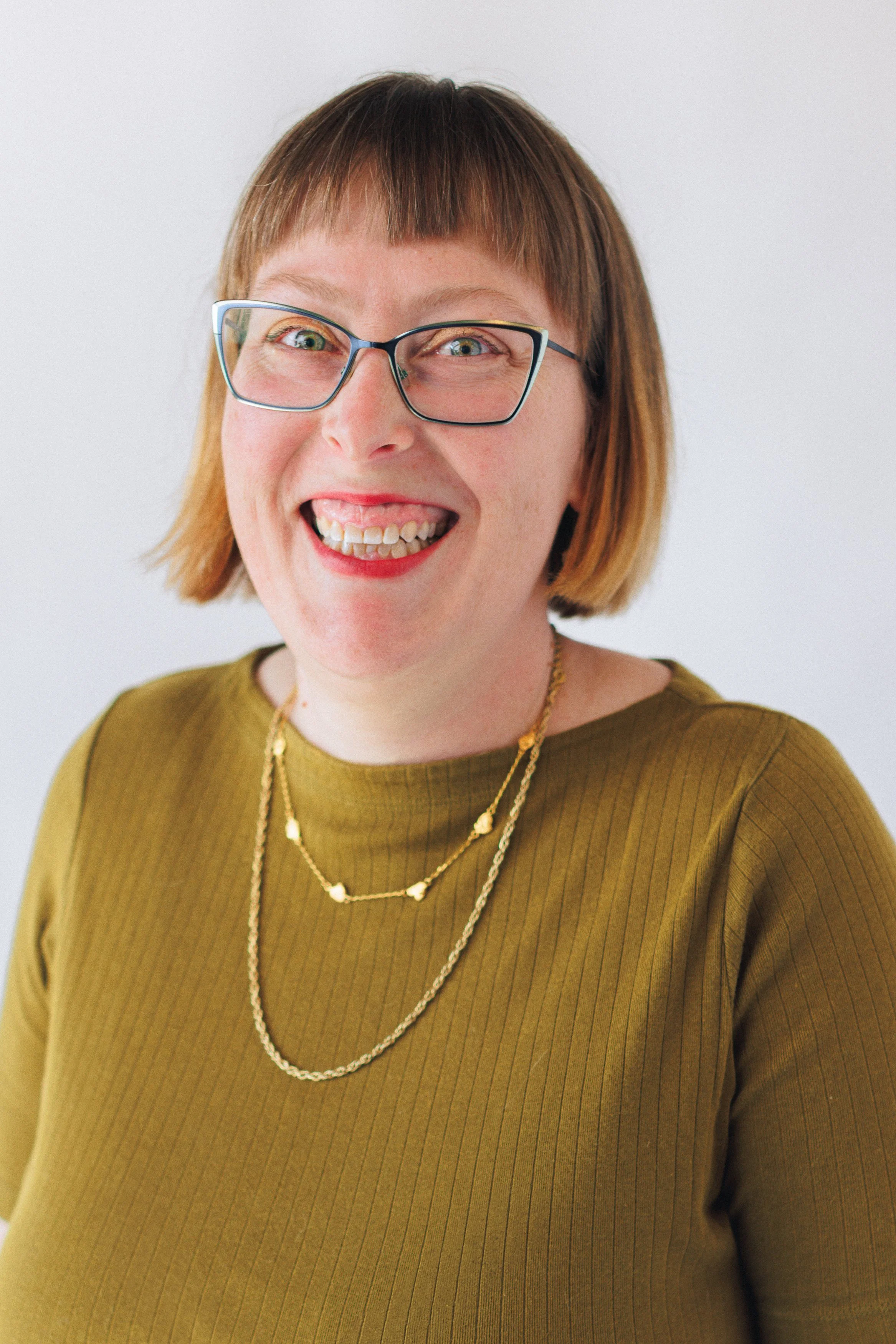 A woman with short brown hair and glasses smiling at the camera, wearing a mustard green top and gold layered necklaces against a plain white background.