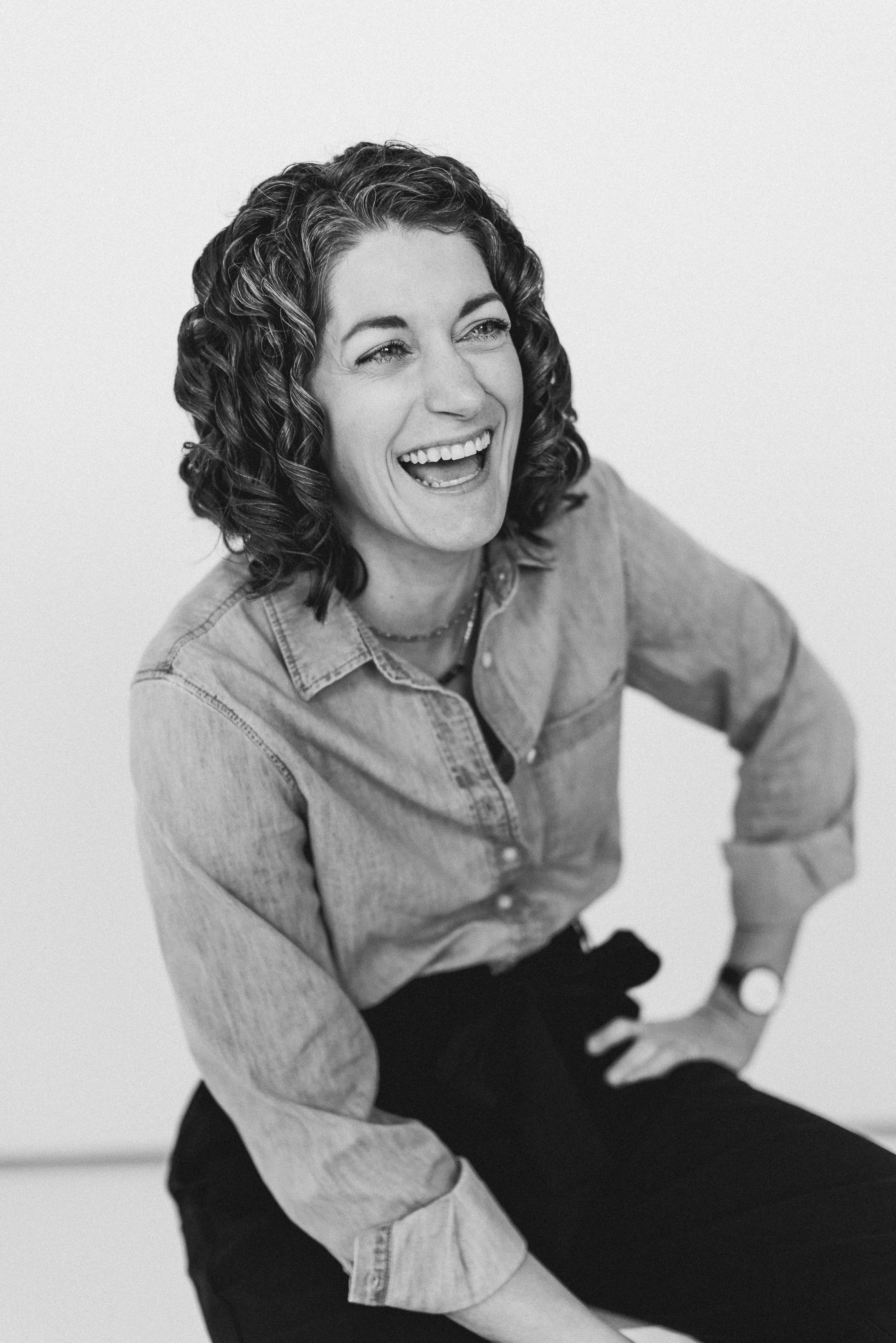 A woman with curly hair laughing, wearing a denim shirt and black pants, sitting against a plain light background.