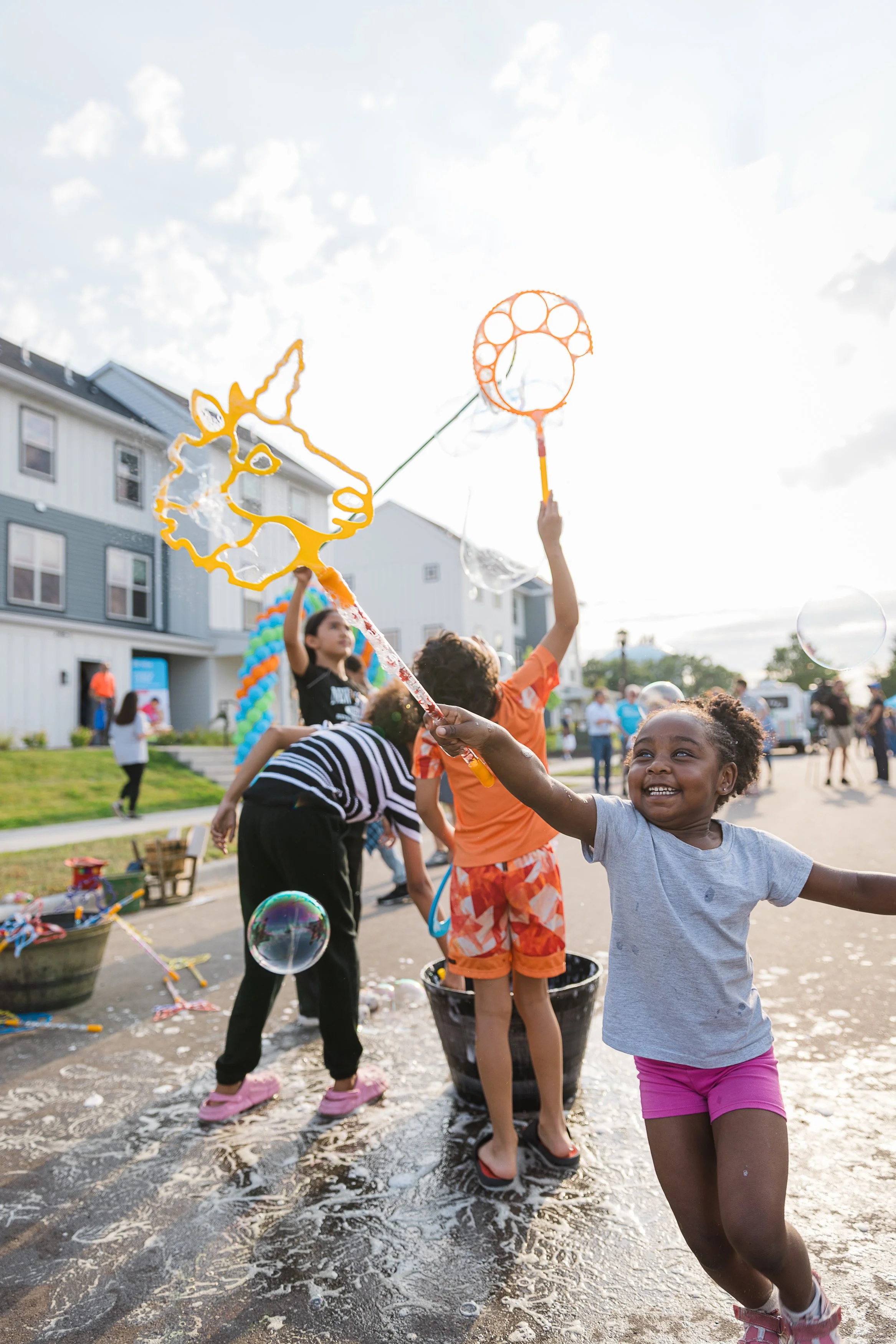 A group of children playing outdoors with bubbles during daytime, some holding bubble wands, on a sunny day with clouds, on a city street with buildings and colorful balloons in the background.