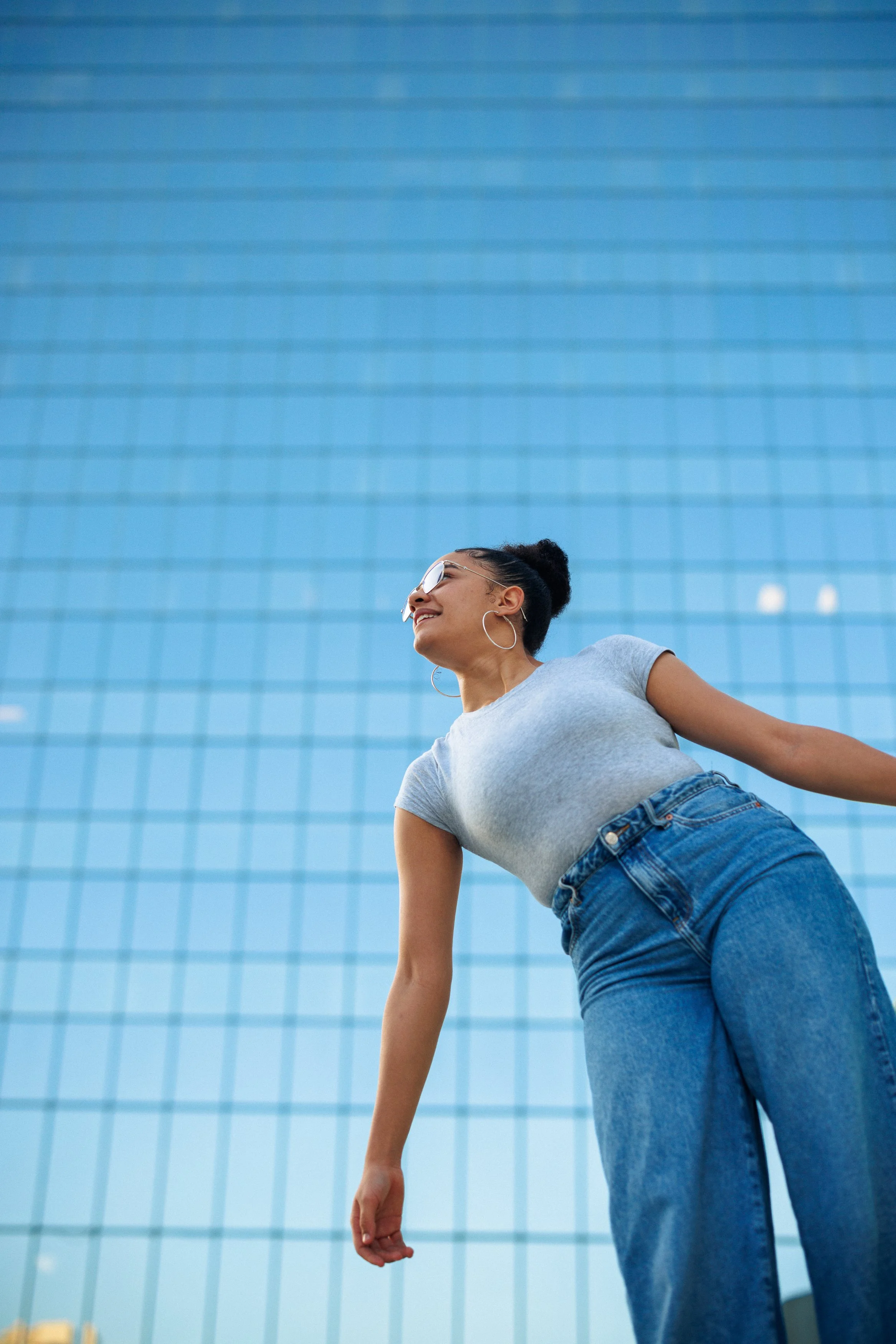 A woman with black hair in a bun, sunglasses, hoop earrings, grey T-shirt, and blue jeans smiling and leaning back in front of a tall glass building.