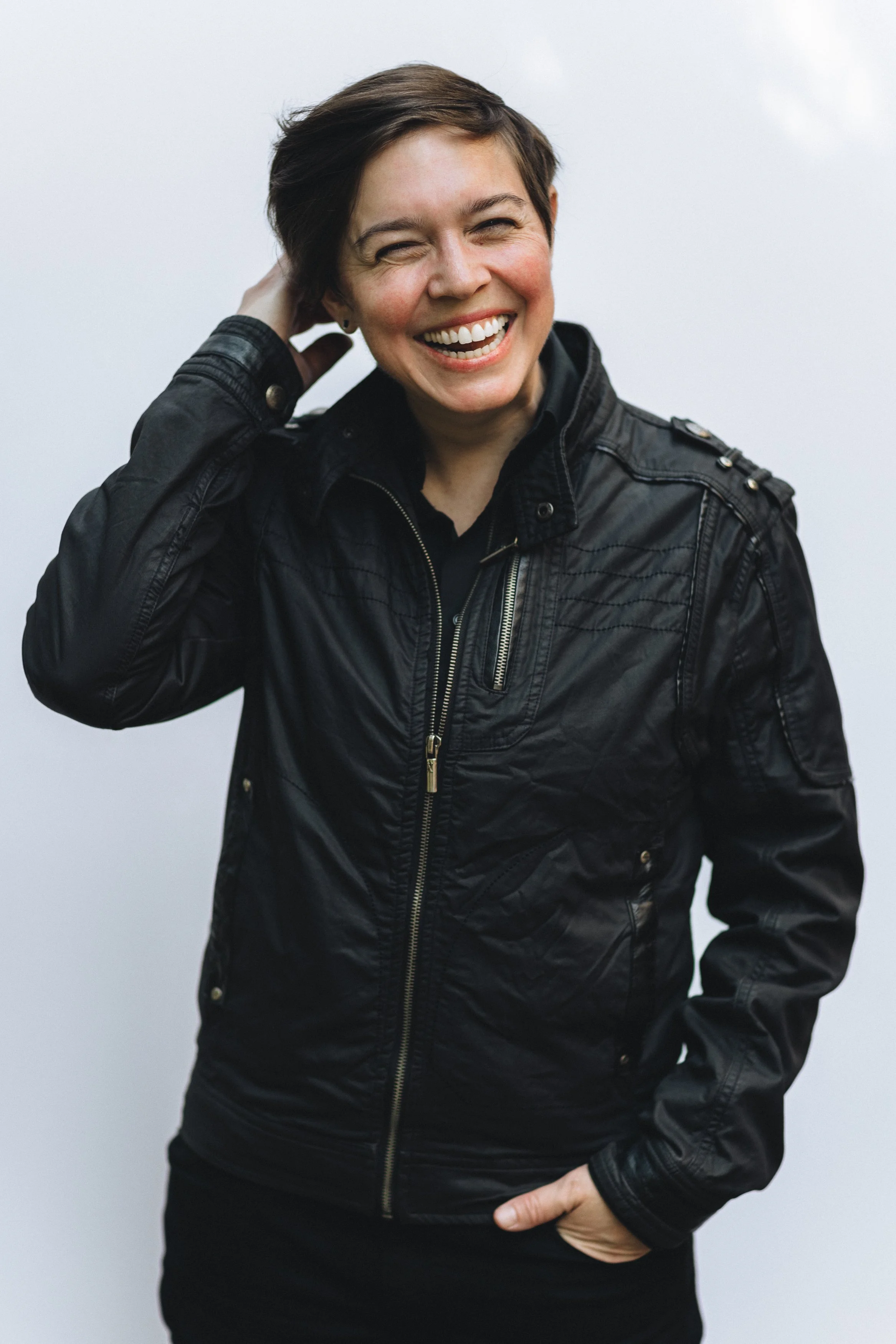 A woman with short brown hair, smiling, wearing a black leather jacket, standing against a light background.