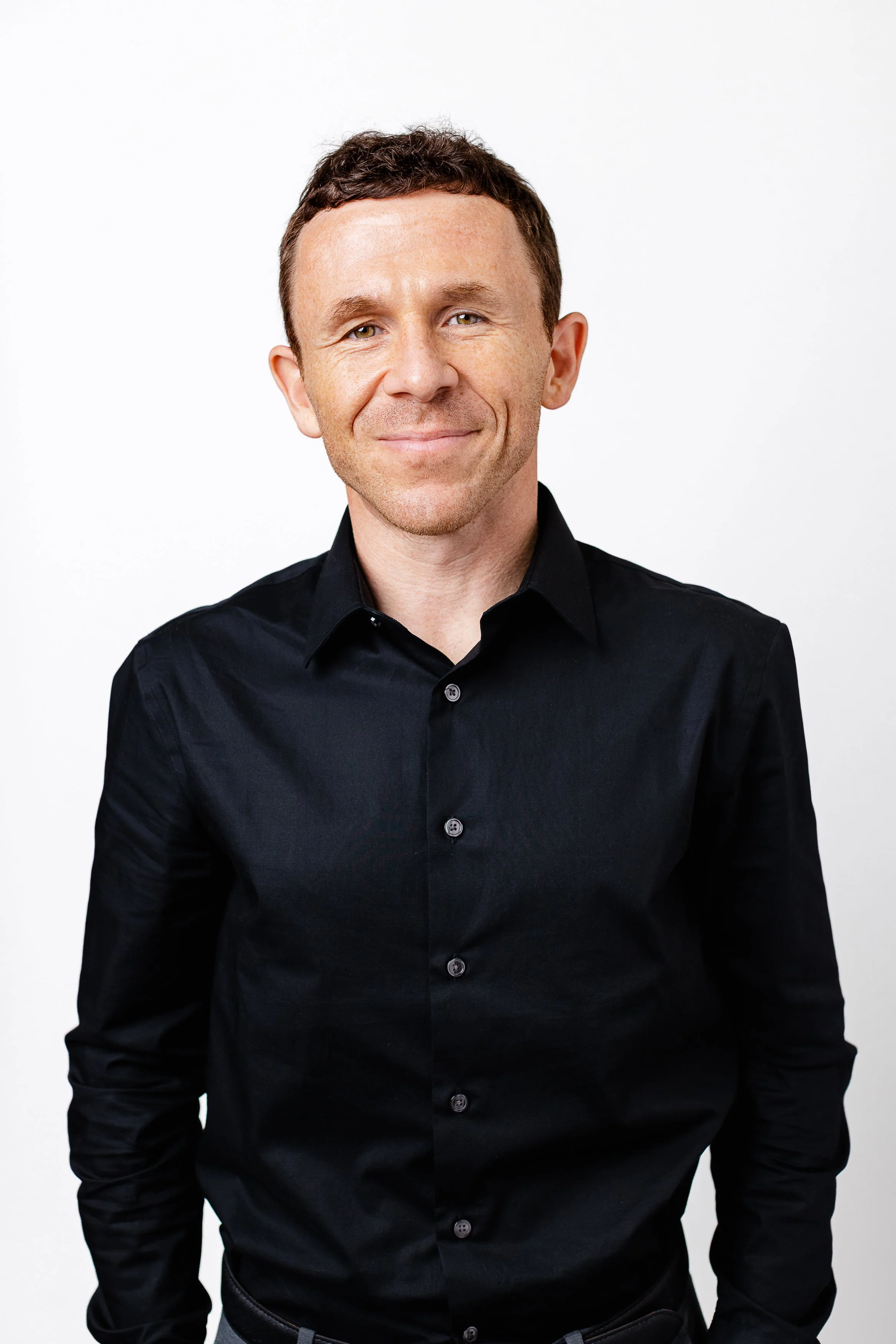 Portrait of a smiling man with short dark hair wearing a black button-up shirt against a plain white background.