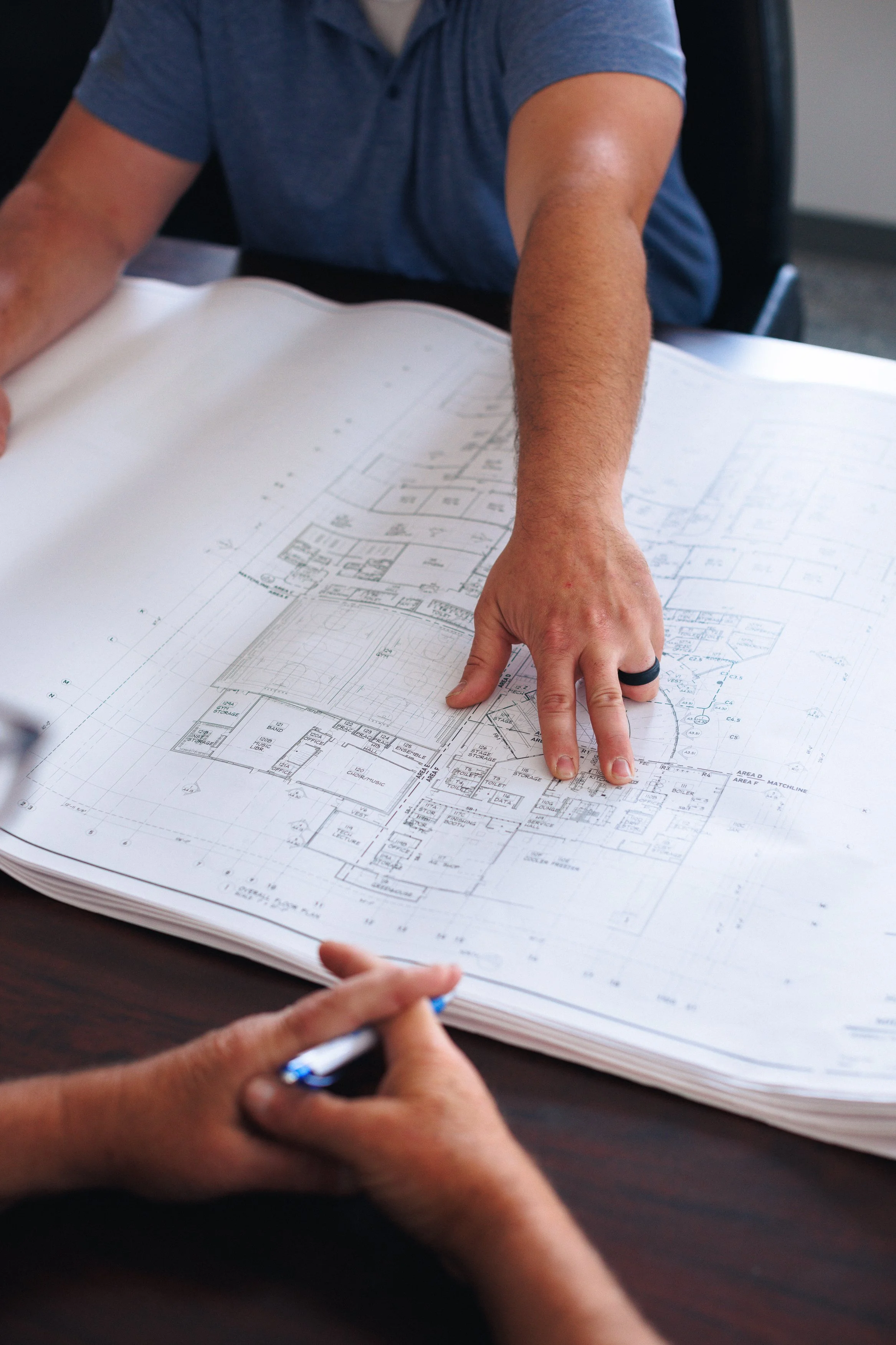 Person pointing at architectural blueprints on a table, with another person holding a pen.