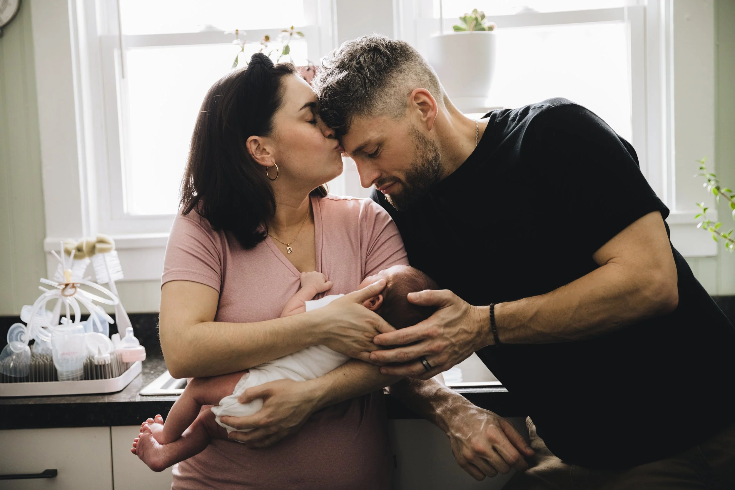 A woman holding a newborn baby while a man kisses her forehead in a kitchen with natural light.