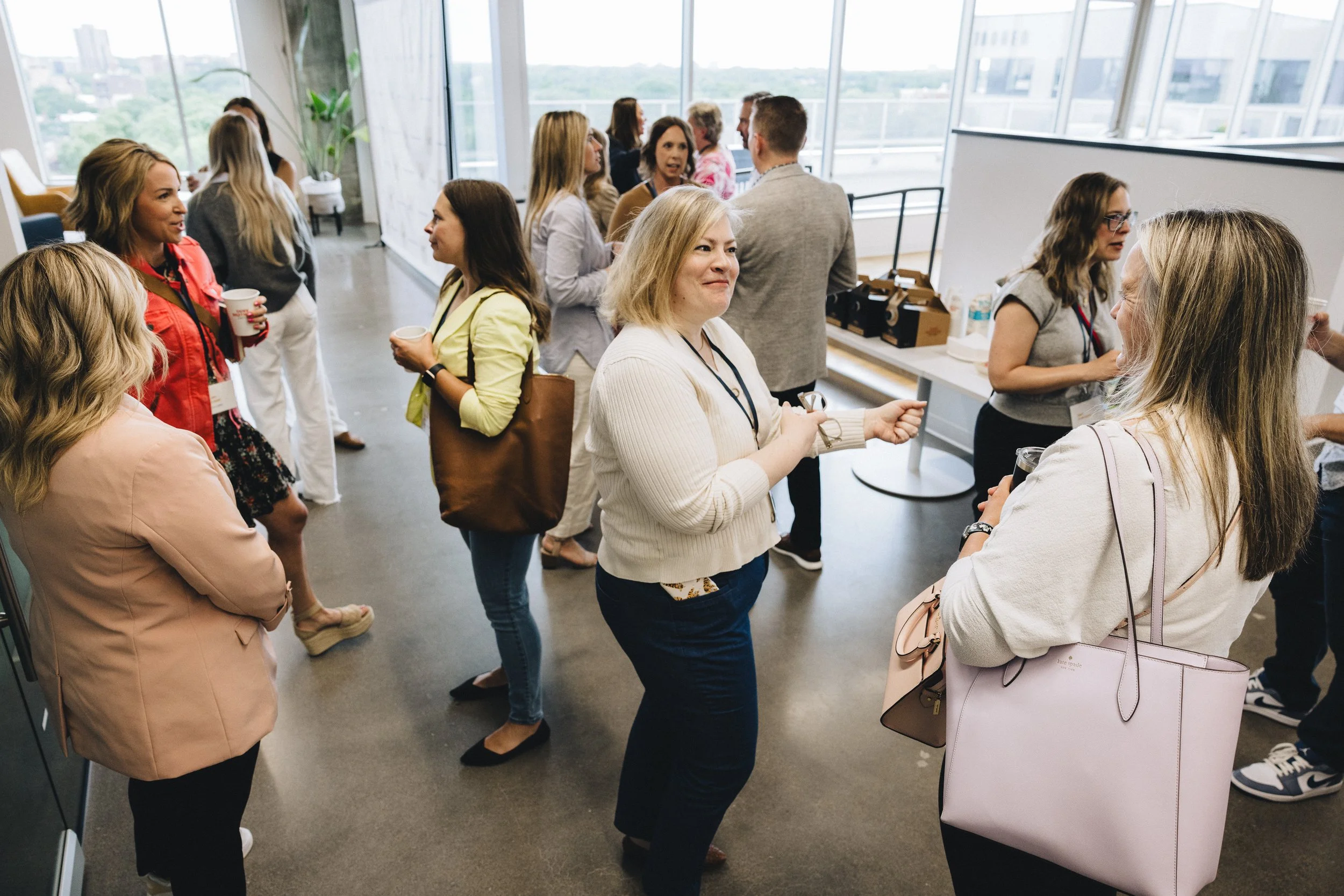 Group of people at a professional networking event or conference, engaging in conversations and holding drinks in a well-lit room with large windows.