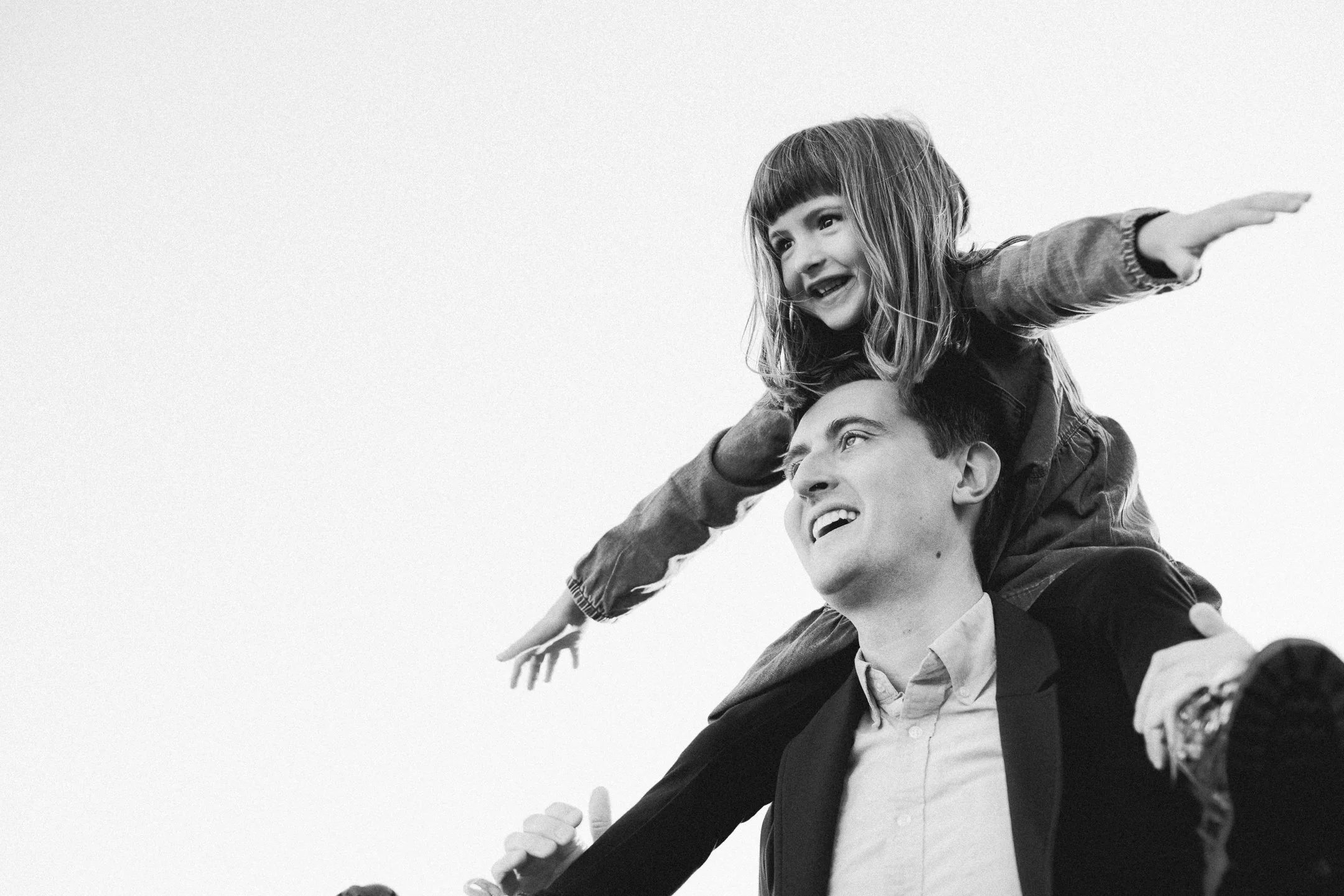 A man carrying a young girl on his shoulders; she has her arms outstretched and is smiling, while he looks up with a happy expression.