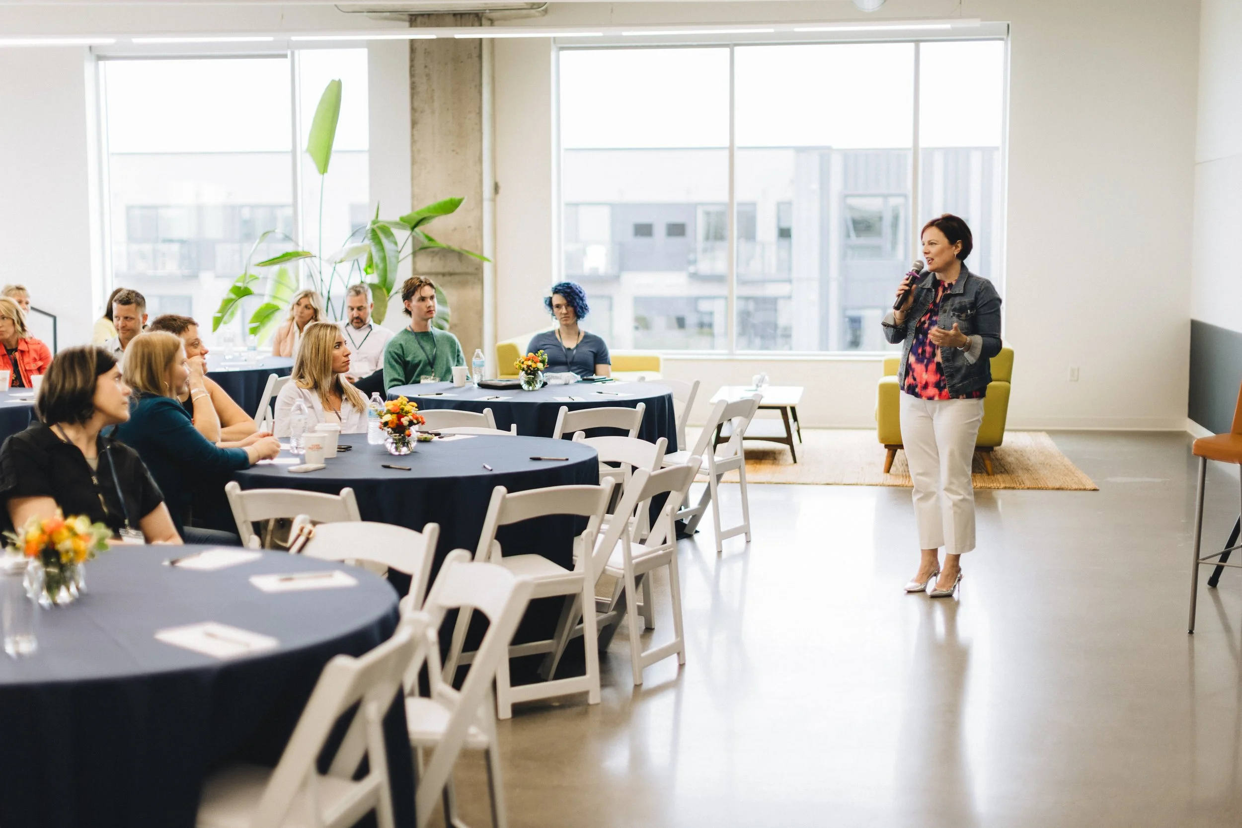 A woman stands in front of a group of people seated at round tables, holding a microphone and speaking in a bright, modern conference room with large windows and minimal decor.